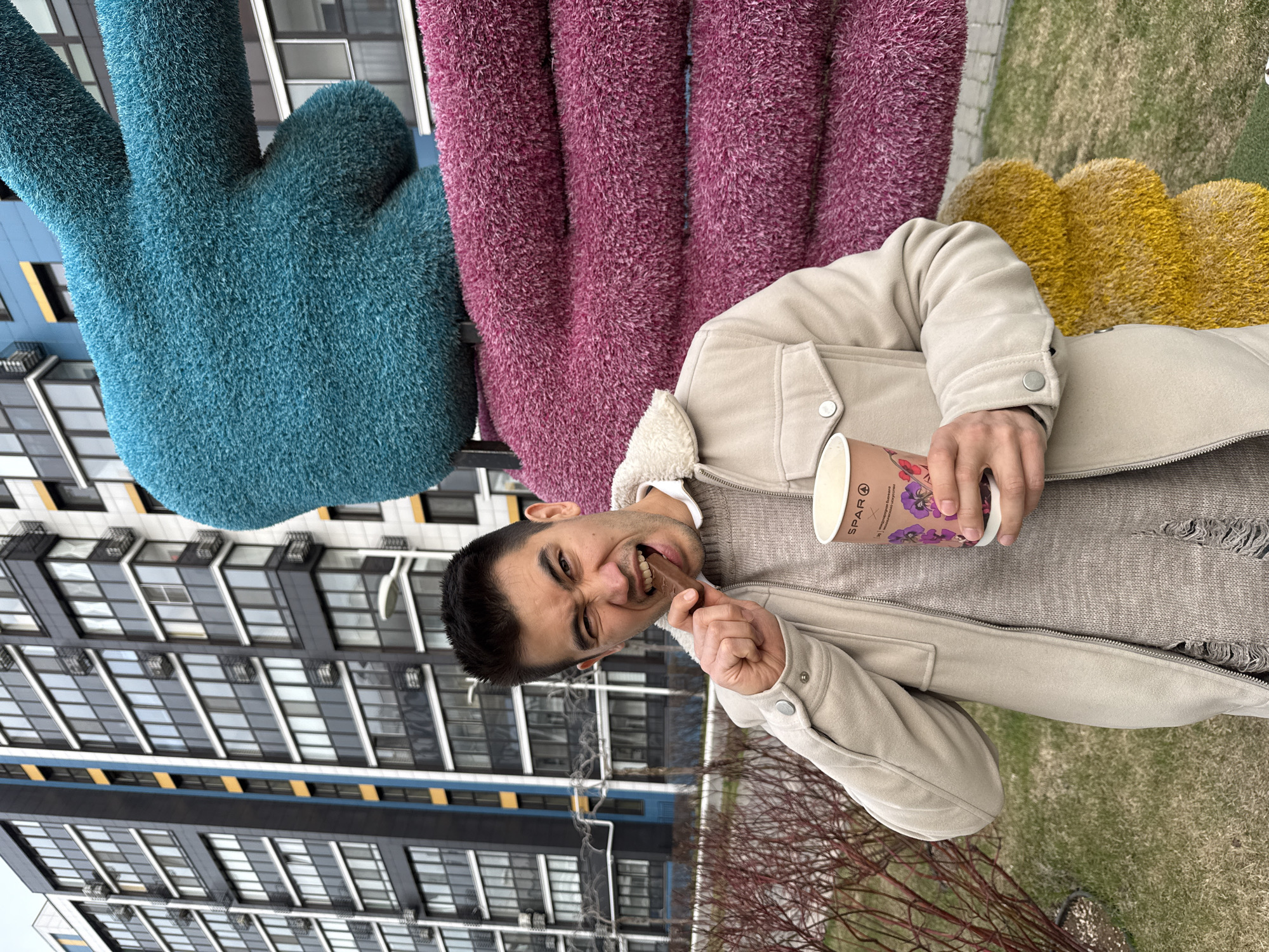 An outdoor, slightly rotated portrait captures a young man in Kazan, Russia, playfully biting into a chocolate bar while holding a coffee cup. He has dark hair and a light complexion, looking towards the viewer with an expressive, almost exaggeratedly joyful or mischievous grimace as he bites the chocolate. He is dressed in a light beige, possibly shearling-lined jacket over a textured, light brown or grey knitted sweater.
In his left hand, he holds a dark chocolate bar, while his right hand holds a white disposable coffee cup. The cup features a colorful floral design, primarily purple pansies, and the visible text "SPAR 4" along with some partial Cyrillic characters.
The background is vibrant and urban, dominated by large, fuzzy outdoor sculptures. Immediately behind the man and to his left is a large, turquoise-blue sculpture shaped like a hand making a peace sign. To his right, there are segments of similar oversized sculptures in deep magenta-purple and yellow. Behind these colorful installations, modern, multi-story residential buildings with numerous windows rise, providing a geometric backdrop. In the foreground, below the man, are bare, reddish-brown branches of bushes and patches of green grass, suggesting an early spring or late autumn setting. The lighting is bright but overcast, indicating daytime. The man's expression conveys a sense of fun and enjoyment.