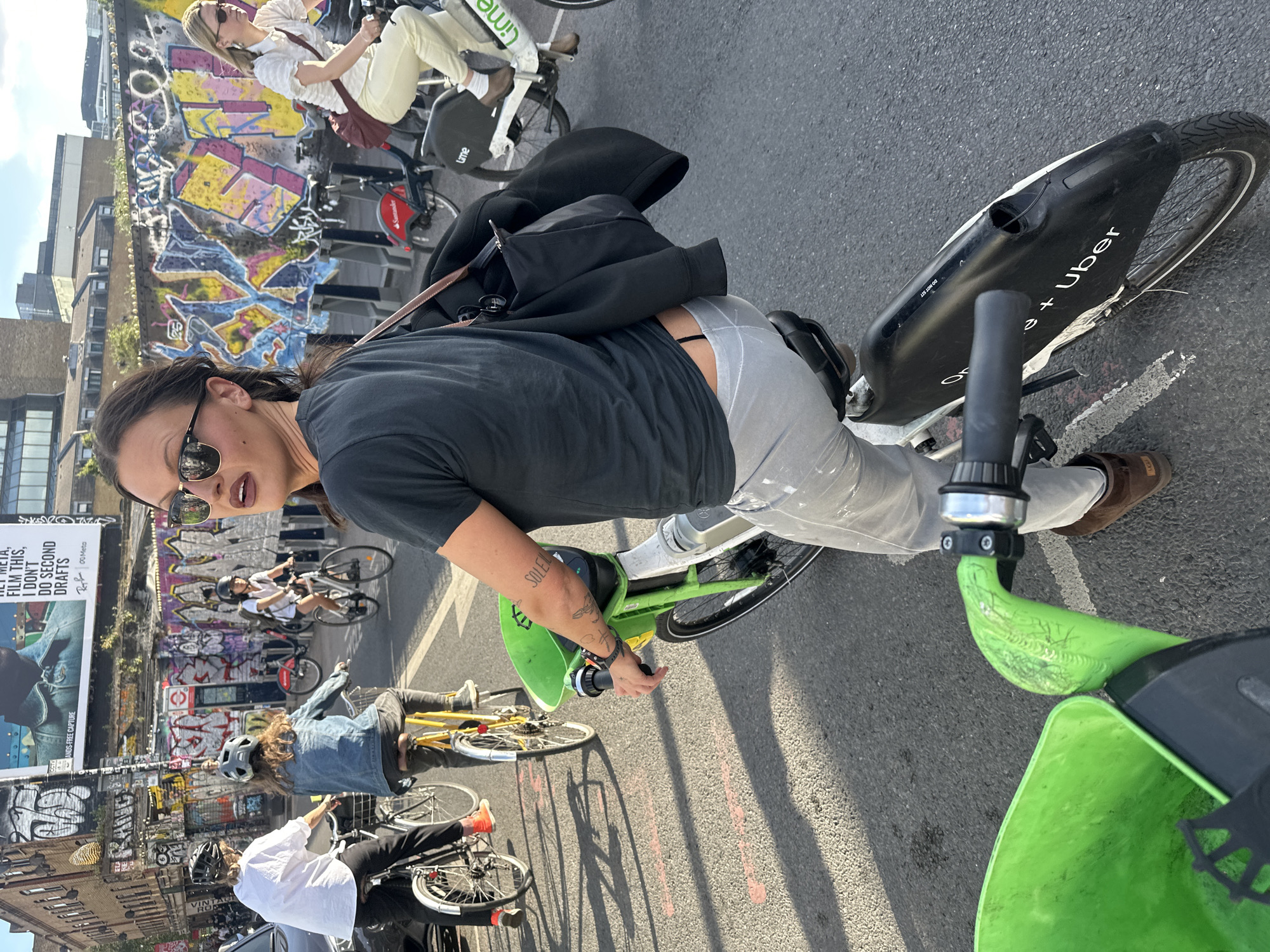 The image depicts a group of people riding bicycles in what appears to be a busy urban environment. One person wearing a black shirt and light grey pants, sunglasses, and a black bag over the shoulder is on a green bicycle and looking toward the camera. There's a tattoo on their arm. Another person in a white shirt and light pants is also riding a bicycle in the background, and a group of people are biking behind them. The bicycles appear to be docked and unlocked, possibly for rent. There is graffiti on a wall or structure nearby, and a billboard is visible in the background. The bicycles are branded with "Lime" and "Uber", suggesting a bike-sharing service. The billboard mentions "Meta" and "second drafts". Based on the style of graffiti, the bicycles, the billboard, and overall urban setting, it is likely the location is in London, United Kingdom.