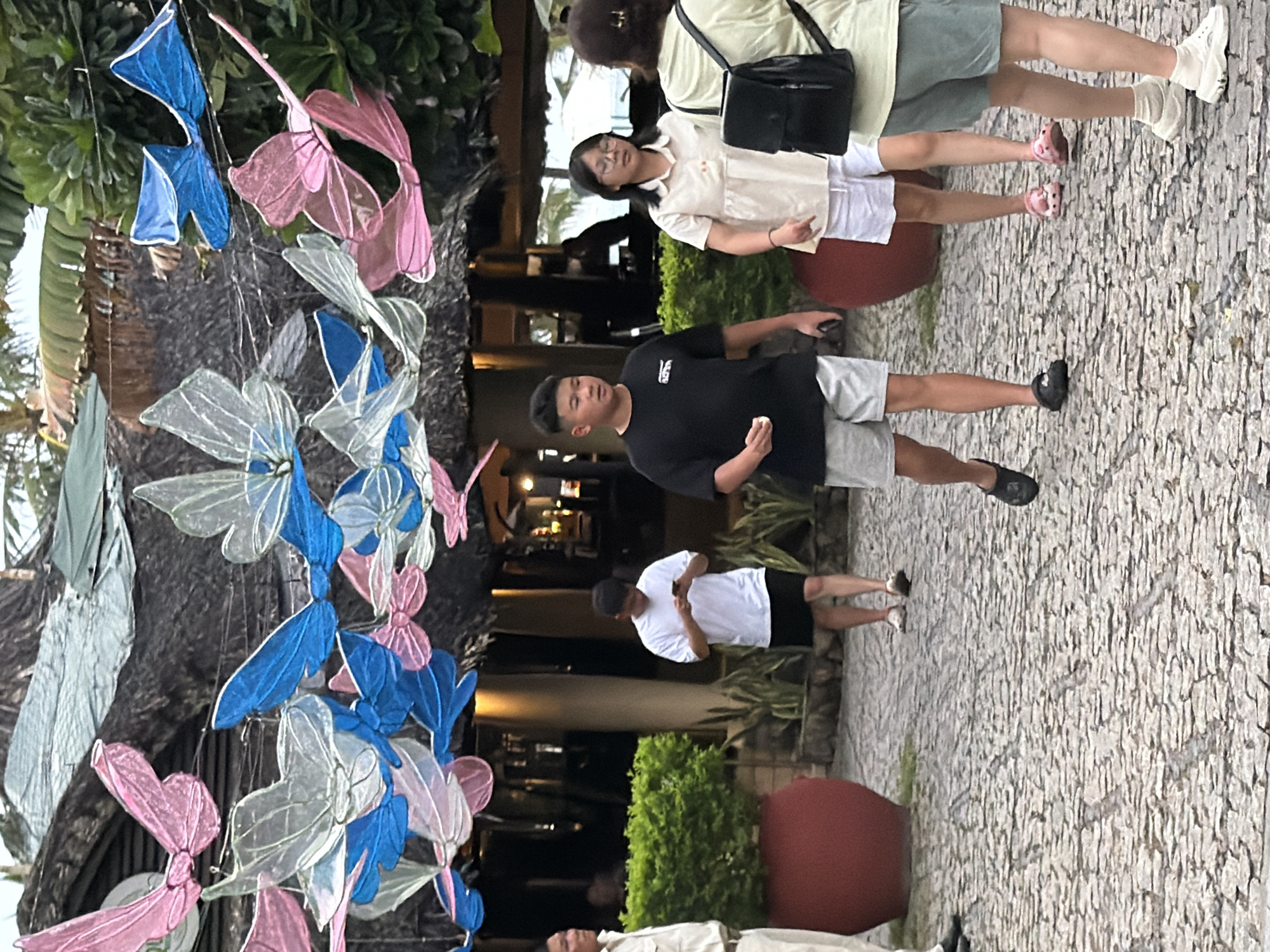 The image depicts an outdoor scene with several people walking on a cobblestone pathway. In the foreground, a young boy in a black t-shirt and grey shorts walks towards the viewer, holding a small white object in his hand. To his right, a girl wearing a light-colored blouse and white shorts is also walking, looking downwards. Further to the right, a person in a light green t-shirt and grey shorts is walking away from the viewer. Behind them, a man in a white t-shirt and black shorts is looking at his phone. On the far left, a man in a light-colored uniform stands facing left. The background features lush green bushes, large red planters, and a building with a thatched roof, adorned with colorful, large butterfly-shaped decorations made of translucent material in pink, blue, and white. The overall setting appears to be a resort or a public area in a tropical location, possibly Nha Trang, Vietnam, given the presence of tropical foliage and the style of architecture.