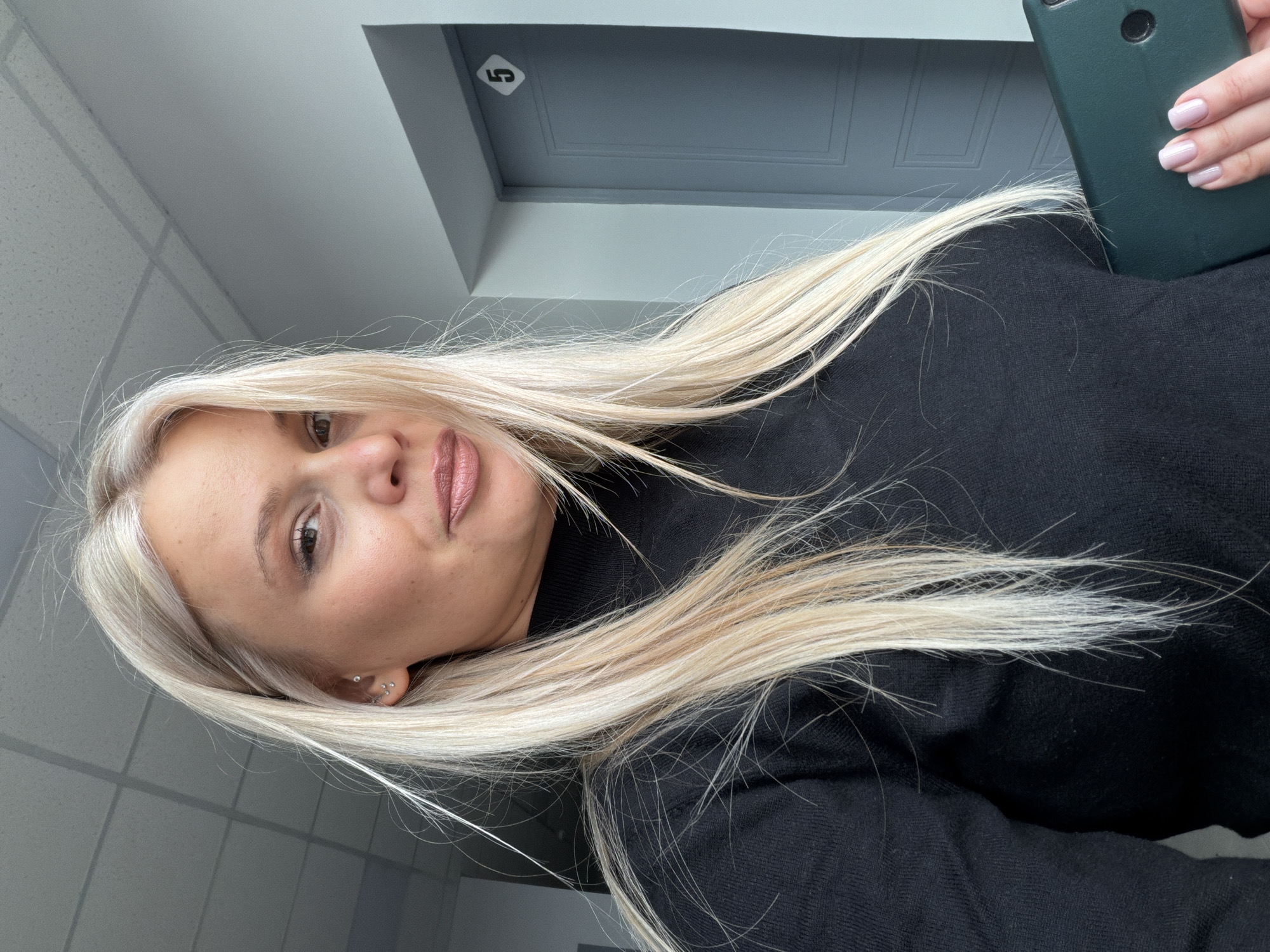A woman with fair skin and blonde hair is holding a green smartphone in her right hand. She is wearing a black turtleneck sweater. Her nails are manicured with light pink polish. The background is an indoor setting with gray walls and a gray door with the number "5" on it. The location is undetermined and there is no verifiable evidence that this image was taken in Sarapul, Russia.