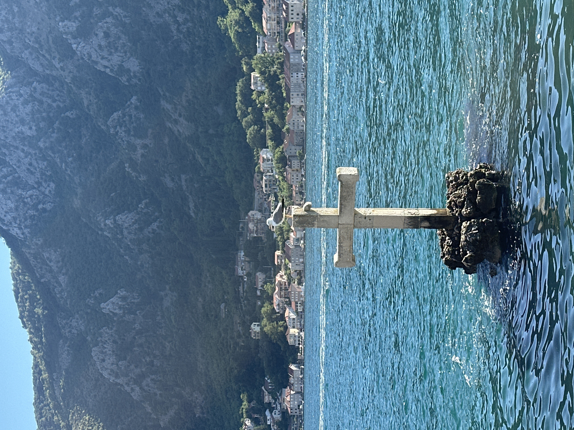 A stone cross stands in the bright blue waters of the Bay of Kotor, in the town of Dobrota, Montenegro. The cross is built on a small cluster of rocks and is adorned with a solitary seagull perched on its top. In the background, the lush green hills of the bay rise steeply, dotted with the terracotta roofs of houses that cascade down towards the water. The sun is shining, casting a warm glow over the scene and creating a rippling effect on the water's surface. The overall impression is one of peaceful serenity, with the cross acting as a silent guardian over the picturesque bay.
