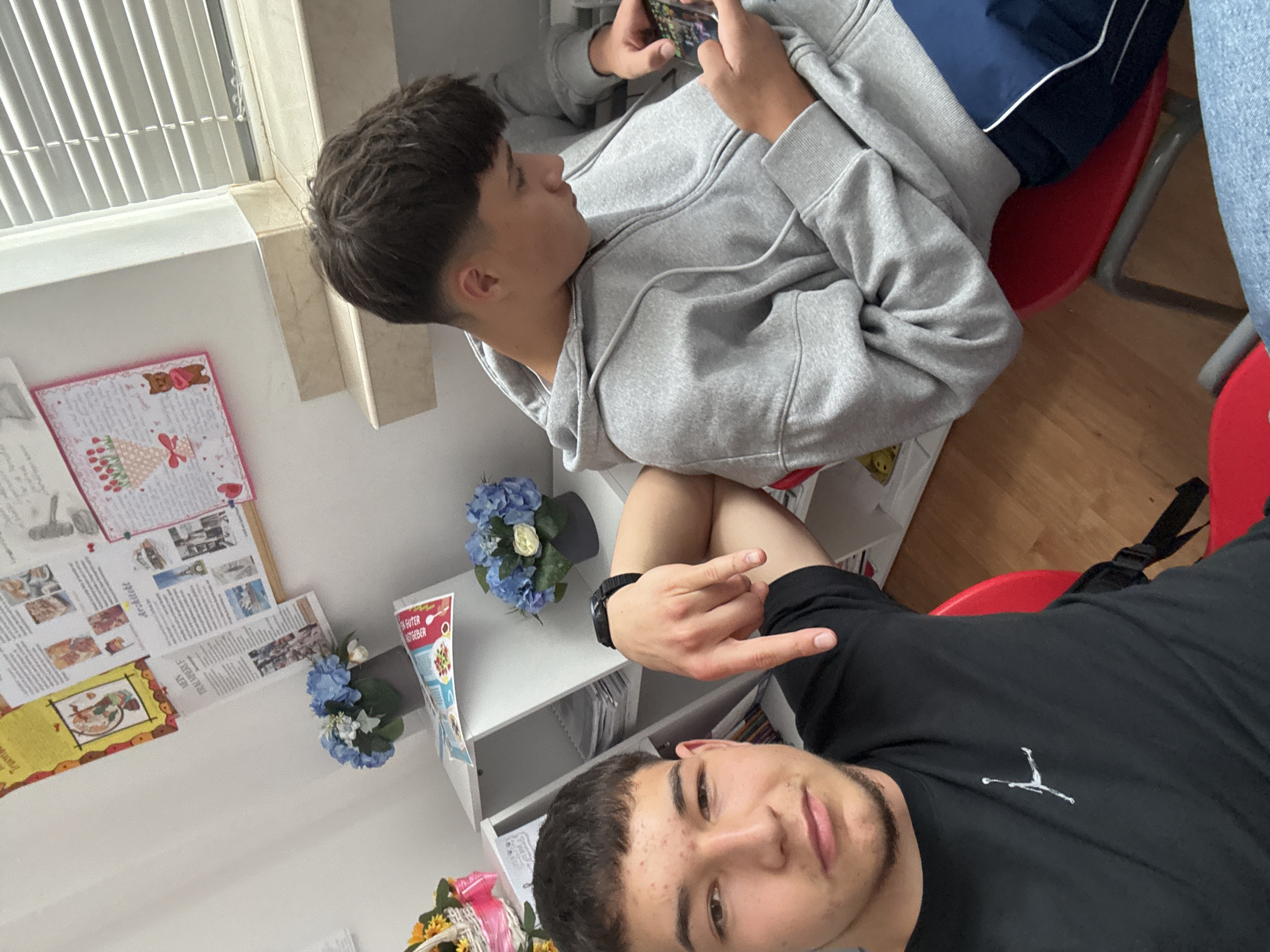 Two young men are indoors, possibly in a school or educational setting, judging by the bulletin boards and student work visible on the wall. One man, wearing a light gray hoodie and dark blue athletic shorts, is seated and using a smartphone. The second man, wearing a black t-shirt with a white Jumpman logo, is positioned closer to the camera and making a hand gesture. There are also red chairs and artificial flowers on a white shelf unit. The photo may have been taken in or around the city of Zlatograd, Bulgaria.