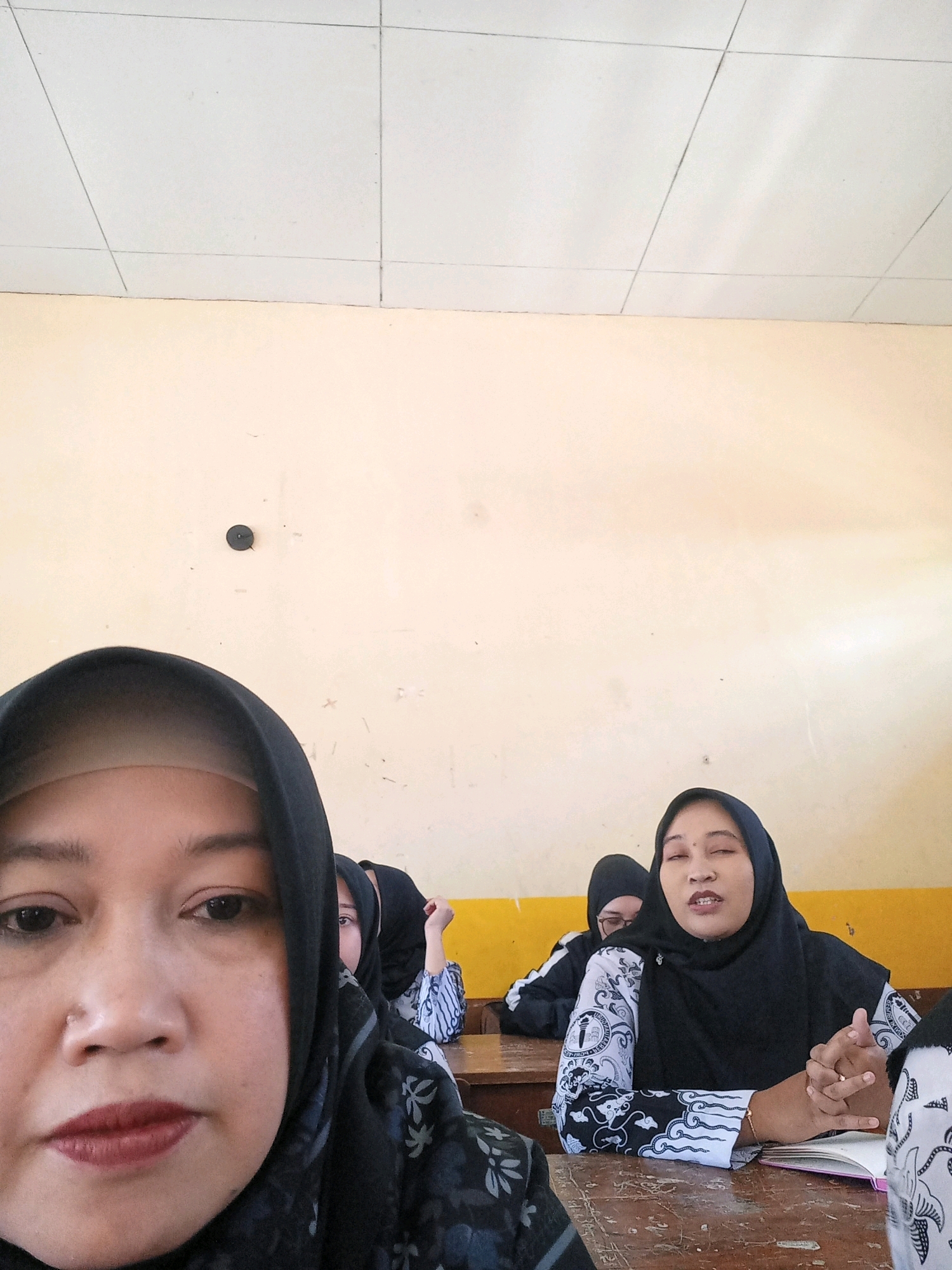 A medium shot captures a group of individuals, likely women, in what appears to be a classroom or meeting room setting. The foreground is dominated by the close-up of a woman's face, wearing a black hijab and part of a black and grey patterned garment. Her eyes are open, and her lips are painted with a deep red lipstick. In the middle ground, another woman in a black hijab is seated at a wooden desk, her eyes are closed, and she is speaking, gesturing with her hands. She is wearing a black and white patterned top. Behind her, another person in a black hijab is visible. The background consists of a light yellow wall with some markings, and a white ceiling with grid lines. The lighting suggests an indoor environment, possibly during the daytime. The scene is devoid of any specific visual cues that would pinpoint its location to Sangubanyu, Indonesia, or any other city.