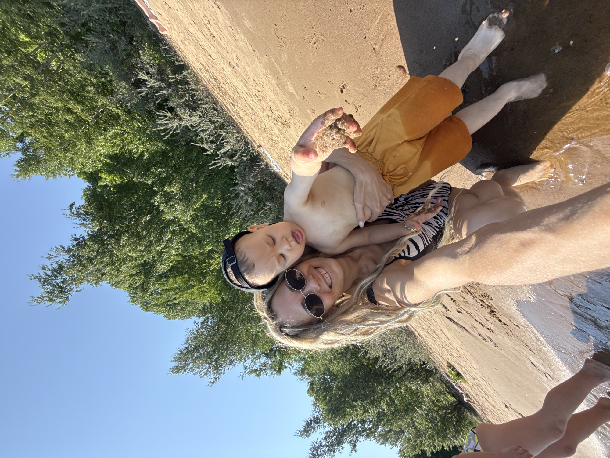 A mother and her young son are posing for a selfie at a beach in Rusanovo, Russia. The mother is wearing sunglasses and a zebra-striped swimsuit. Her long blonde hair is loose. The son is wearing a black baseball cap. He has sand in his hand and is sticking his tongue out. They are standing on the edge of a body of water with their feet in the water. Trees are visible in the background, as well as the sky.