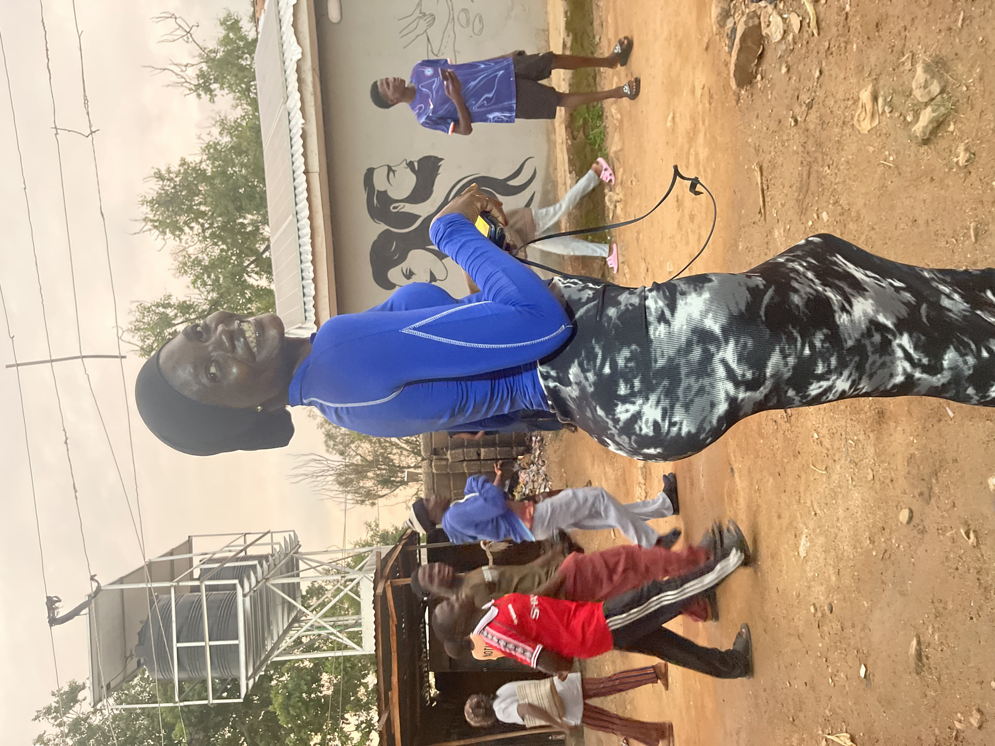 A low-angle, medium shot captures a Black woman wearing a blue long-sleeved shirt and a black and white tie-dye skirt, turning her head to look back at the camera with a smile. She wears a black headscarf and holds a black strap in her right hand. The background depicts a dusty street in the city of Jos, Nigeria, with a metal water tower structure on the left, and a building with solar panels and a mural of a man and woman on the right. Several other Black individuals, appearing to be young men, are walking and standing in the background, some wearing athletic attire. The ground is uneven and dusty, with scattered debris. The sky is overcast and light.