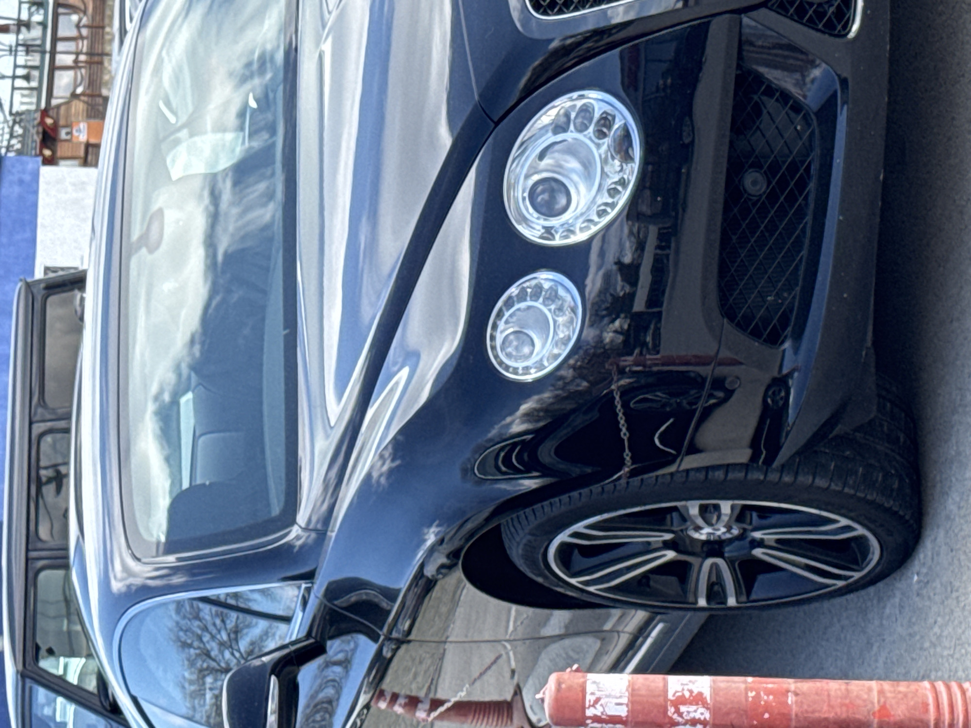 A close-up photograph shows the front of a dark-colored Bentley. The distinctive round headlights and mesh grille are visible, as is part of the front passenger-side tire. The car is situated on an asphalt surface, and an orange and white bollard lies at the bottom of the frame. The reflection in the windshield suggests an outdoor setting with buildings and trees. While exact landmarks aren't distinguishable, based on the general environment, and assuming the image location is as described, it is likely to be a city street scene within Varna, Bulgaria. Additional vehicles are partially visible in the background.