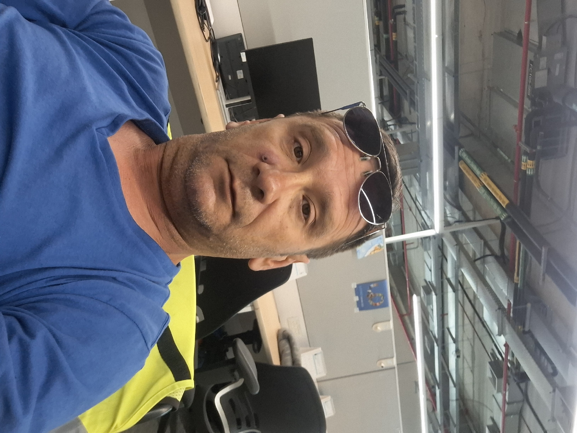 A man is taking a selfie in an office environment. He is wearing a blue t-shirt and sunglasses are resting on top of his head. There is a monitor and office chairs in the background, along with what appears to be overhead lighting and ceiling infrastructure typical of an industrial or large office space. The location of Kiryat Gat, Israel is not discernible from the visual elements in the image.