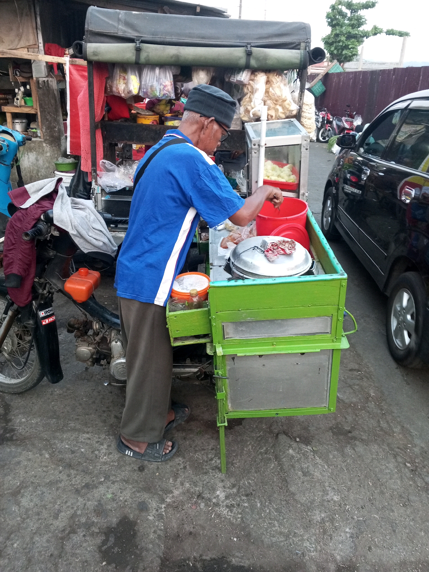 A street vendor in Gentan, Indonesia, stands next to his mobile food cart, which is attached to a motorcycle. The vendor, an elderly man wearing a blue shirt, brown pants, and a black cap, is seen holding a ladle over a red bucket. His cart is a bright green metal structure with a glass display case containing food. Behind him, a black car is parked on the street. The scene depicts a typical street food vendor setup in Indonesia.
