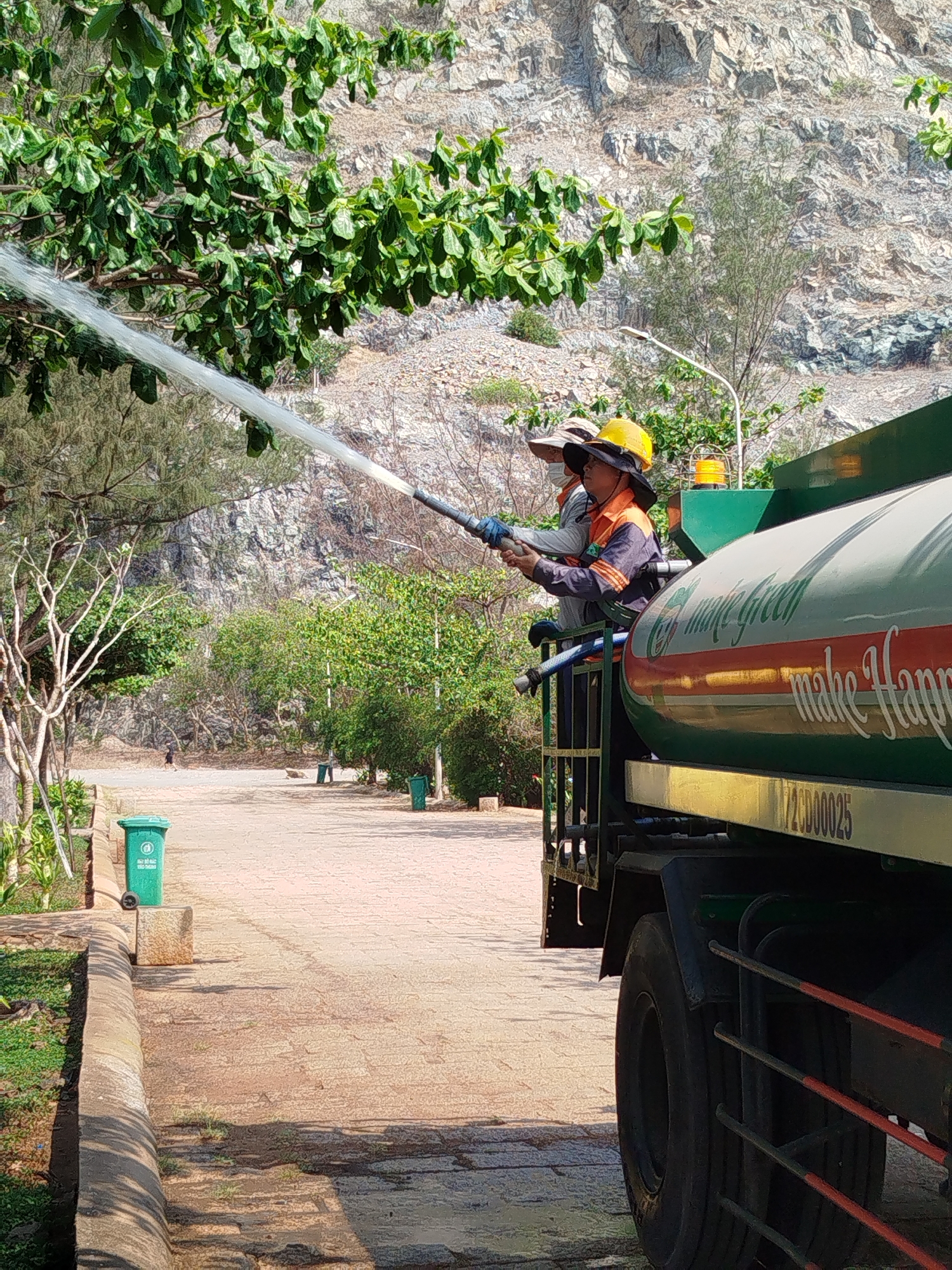 Two sanitation workers in work clothes and hats are on the back of a large green truck. The truck, labeled "make green make Happy", is parked on a paved road, and they are using a hose to spray water into the nearby trees and greenery. In the background is a rocky hillside and various plants. The setting is a park or public space in Ho Chi Minh City, Vietnam, as indicated by the trash bin with Vietnamese writing visible in the foreground.