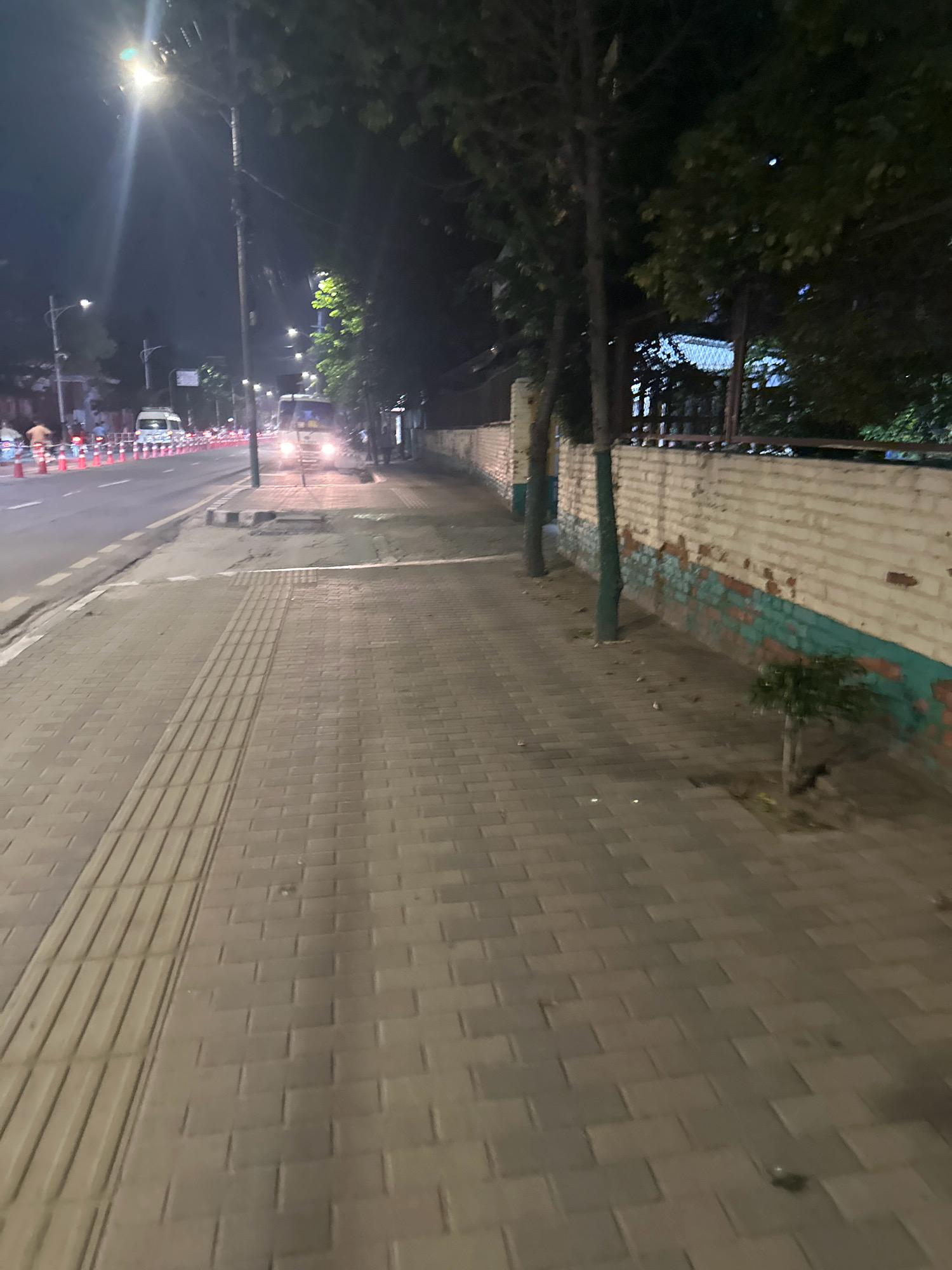 A nighttime street scene in Kathmandu Metropolitan City, Nepal shows a paved sidewalk adjacent to a road. The sidewalk is made of interlocking gray pavers and features a tactile paving strip. The road has traffic cones lined up along its edge and a white van is visible in the distance, its headlights illuminated. Trees line the edge of the sidewalk, casting shadows. A low brick wall with peeling paint runs along the far side of the sidewalk. Streetlights illuminate the scene.