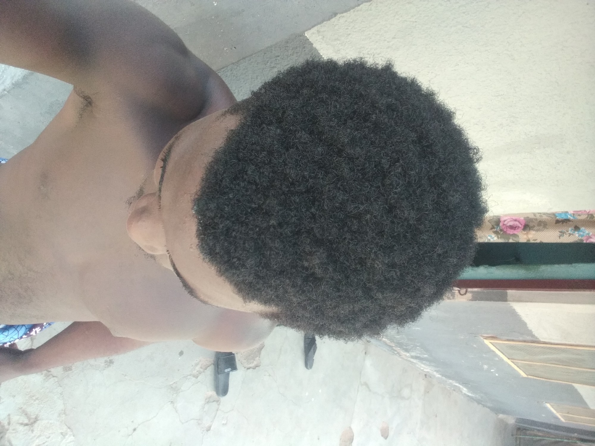 The image is a high-angle, full-body shot of a Black male in Jalingo, Nigeria. He is standing shirtless, facing away from the camera, with his head turned to the side. His hair is dark, curly, and appears to be of medium length. The man is wearing patterned blue and white shorts.
The environment appears to be an outdoor space with a concrete floor. To the left of the man is a lightly textured, beige wall. To his right, there is a building with a dark gray painted wall and windows with light-colored frames. A floral curtain is visible on the left side of the frame, partially obscuring a doorway that leads into a room with a turquoise-colored interior. Two pairs of dark flip-flops are visible on the concrete floor behind the man. The lighting suggests it is daytime.