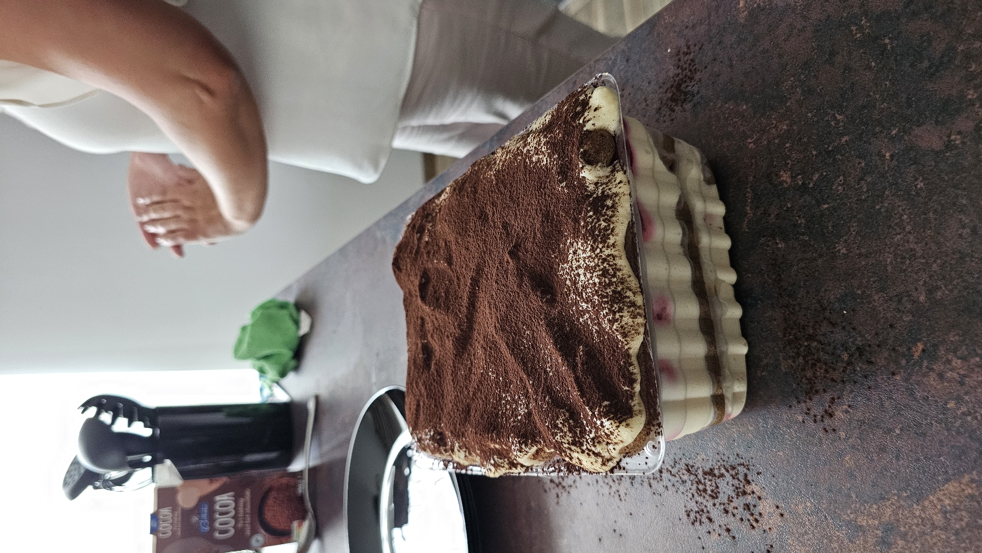 The image depicts a close-up, eye-level view of a rectangular tiramisu dessert in a clear plastic container, dusted heavily with cocoa powder. The dessert has visible layers of cream and what appear to be soaked ladyfingers, with hints of pink fruit visible between the layers. The tiramisu is placed on a dark, textured countertop. To the left of the dessert, a black utensil holder with several kitchen utensils is visible, along with a box of "COCOA" powder. A green cloth is also present on the countertop. In the background, a person wearing a white top and light-colored pants is partially visible, with their hands clasped. The environment suggests an indoor setting, likely a kitchen. The image does not provide any information about the city of Varna, Bulgaria.