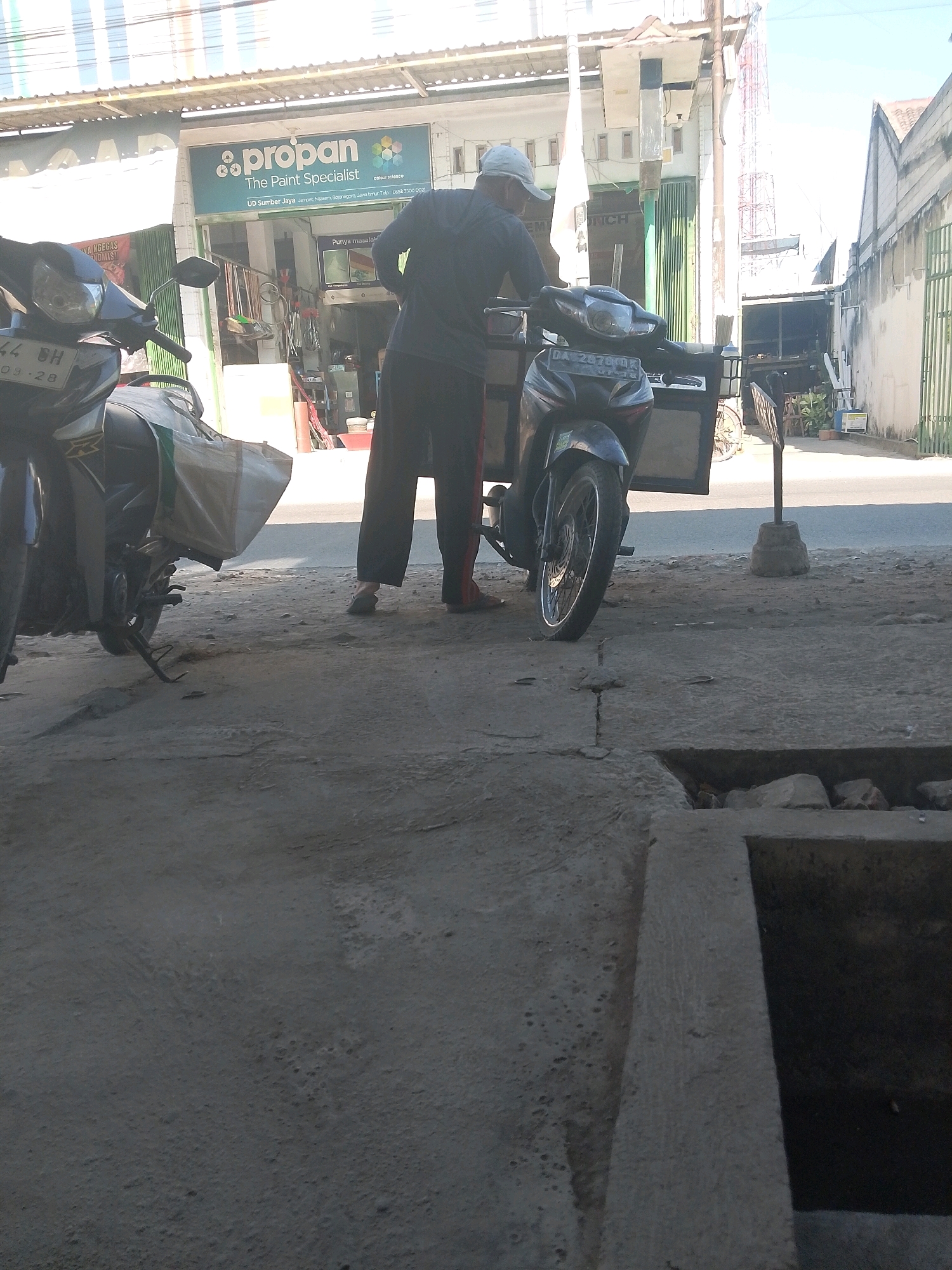In Jampet, Indonesia, a man wearing a light-colored baseball cap and dark clothing, including long pants, stands next to a parked motorcycle. The motorcycle is black and has a cargo box attached to its rear, carrying a white bag. The man appears to be leaning forward slightly, possibly interacting with the motorcycle or its cargo. To the left of the man and motorcycle, another motorcycle, also black, is partially visible. Behind the man and motorcycles is a building with a sign that reads "propon The Paint Specialist." The building appears to be a shop or business. The scene is set on a paved street or sidewalk, with a concrete drain visible in the foreground. The overall environment suggests an outdoor commercial area in a town or city.