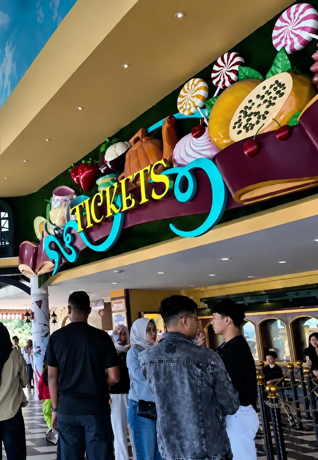 The image depicts the entrance area of a theme park or attraction in Berastagi, Indonesia, featuring a brightly decorated sign that reads "TICKETS" in yellow lettering, surrounded by colorful, oversized candy and fruit-themed decorations. The sign is mounted on a green background and set against a yellow awning.
In the foreground, a group of people are visible. Closest to the camera, a man in a black t-shirt and jeans is seen from behind, facing away from the viewer and towards the entrance. To his right, a woman wearing a light blue hijab and glasses is looking towards the man. Beside her, a person in a grey denim jacket is engaged in a conversation with another individual wearing a black shirt and white pants, also seen from behind. Several other individuals are further back in the queue, including a person in a black hijab and a person in a beige jacket. The environment suggests an indoor or covered outdoor space, with pillars and decorative elements visible in the background. The overall atmosphere appears to be that of an entertainment venue entrance, with people waiting to purchase tickets.