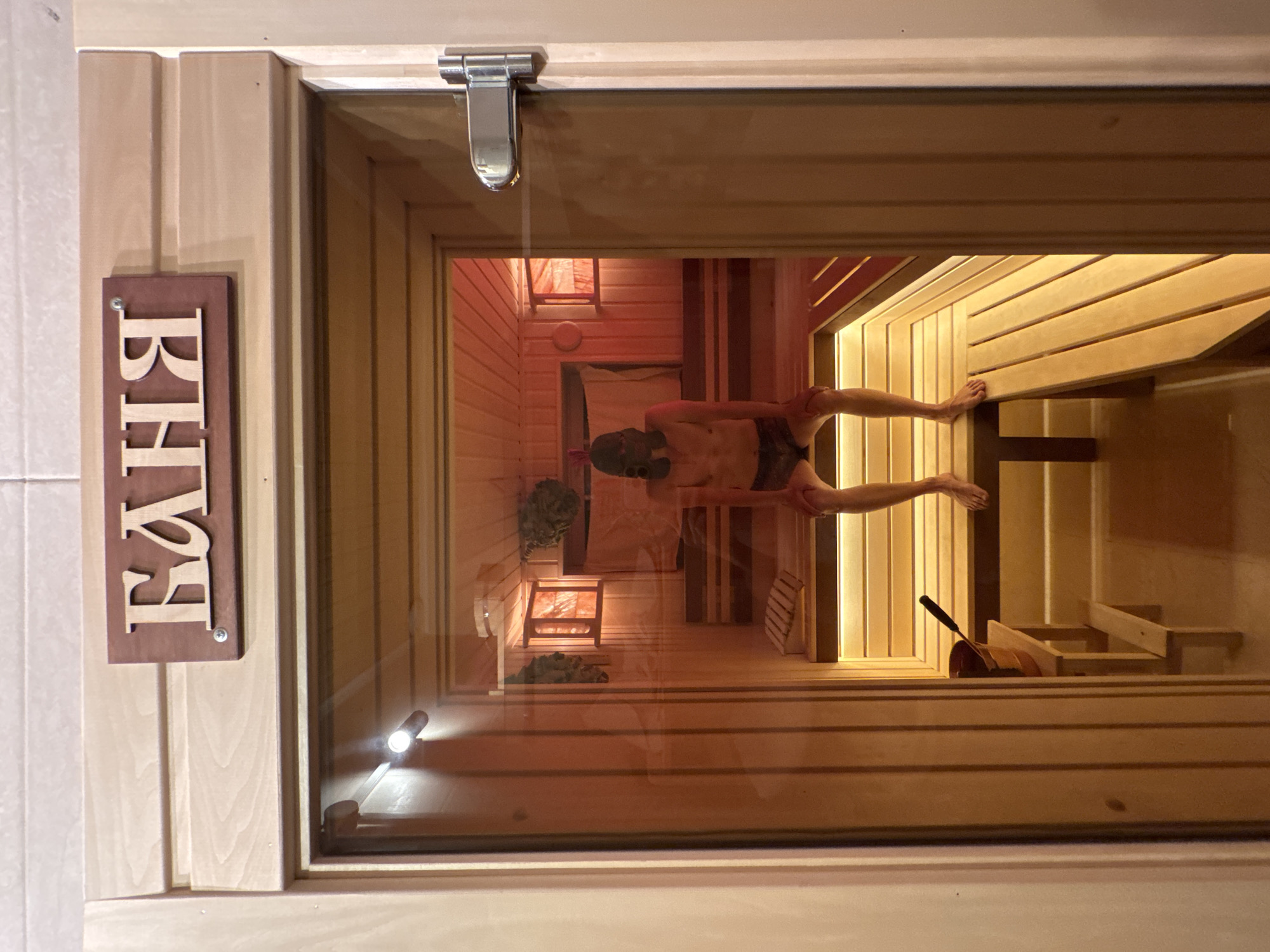 This is a medium shot of a person sitting in a wooden sauna. The sauna entrance is framed by light-colored wood and a glass door. Above the entrance, a wooden sign with Cyrillic lettering reads "БАHЯ" (Banya), which translates to "bathhouse" or "sauna." The interior of the sauna is illuminated by red lights, creating a warm, orange glow. The walls and benches are made of polished wood. The person, who appears to be a man, is wearing dark swim trunks and a dark helmet or head covering. He is sitting on a wooden bench with his knees bent and hands resting on them. To his left, on a small wooden stool, is a bucket with a ladle. In the background, there are bundles of dried branches, commonly known as veniks, hanging on the wall. The floor of the sauna is also made of wood. The scene is set in a sauna located in Zelenaya Roshcha, Russia.
