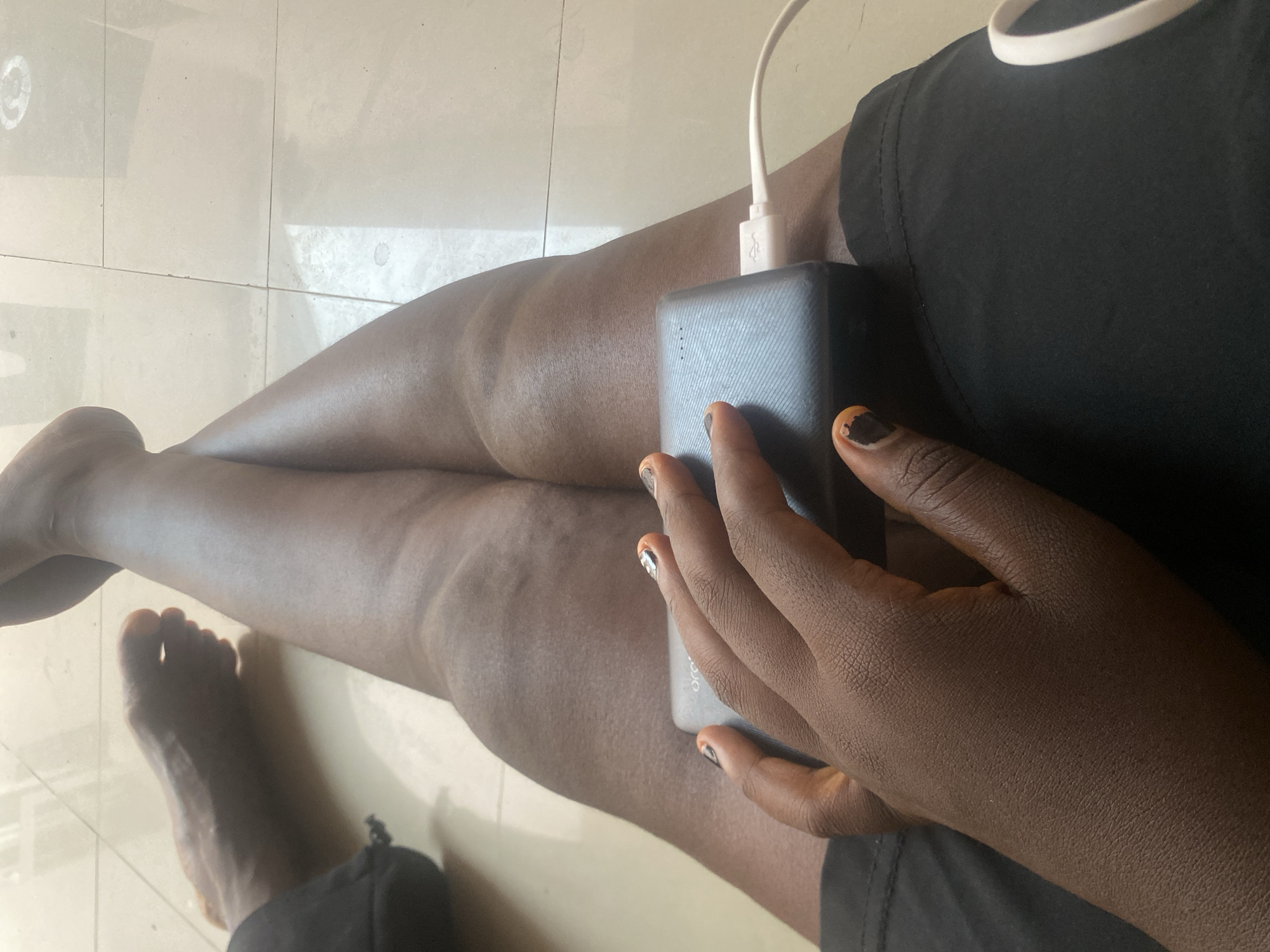 This media file captures a close-up view of a person's legs and hands, set against a tiled floor. The person is wearing a black top and appears to be holding a dark-colored power bank with a white charging cable connected to it. The person's nails are painted with a distinct design, featuring orange and black colors. Visible on the power bank is some faint text, though it is largely illegible. The lighting suggests it is daytime, and the setting is likely indoors, given the tiled flooring. The image is framed to focus on the person's legs and the power bank, with one foot also visible. The overall scene is casual and everyday.