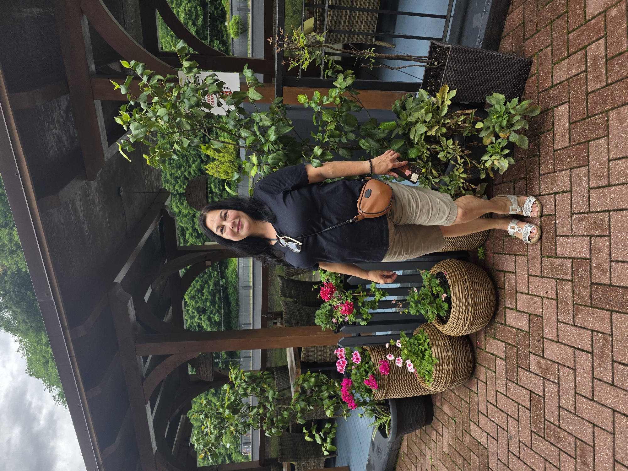 A woman with long dark hair stands on a brick patio beneath a wooden covered structure. She is wearing a dark t-shirt, light-colored shorts, and silver sandals. A small brown purse is slung across her body. To her left are several potted plants, including some with pink flowers. To her right, a large green plant is in a dark planter. In the background, tables and chairs are visible through the wooden structure, and beyond that, a body of water and lush green trees. The scene appears to be an outdoor seating area, possibly a restaurant or cafe, in Estosadok, Russia, given the presence of Russian text on a sign hanging from the structure.