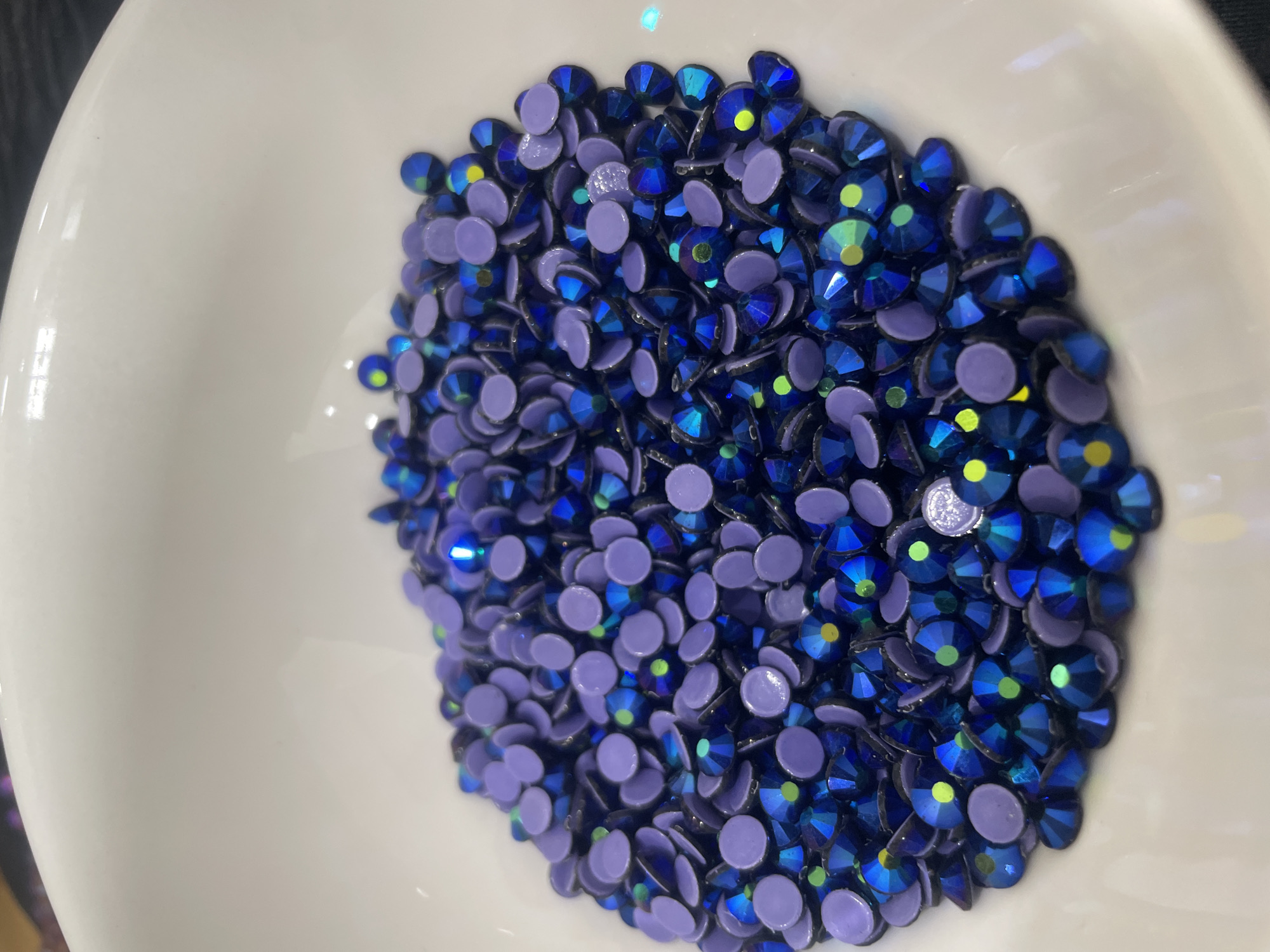 The image presents a close-up, overhead view of a collection of small, sparkling rhinestones or gems resting in a white, shallow bowl. The bowl, which has a glossy finish, occupies most of the frame, with a dark, out-of-focus background visible along the edges, suggesting an indoor setting.
The gems are primarily of two types, intermingled throughout the pile. The most prominent are faceted, dark blue to violet gems that exhibit a strong iridescent or "Aurora Borealis" (AB) effect. These gems reflect light with shimmering hues of green, yellow, and lighter blue, adding a dynamic, colorful sparkle. Scattered among these are numerous opaque, flat-backed, circular gems of a consistent lavender or light purple color, providing a softer contrast to the highly reflective blue ones. Many of the gems have their flat, adhesive-ready backs facing upwards.
The gems are gathered in a dense, irregular pile within the center of the bowl. There are no people or animals visible, and no specific activity is taking place; the gems appear to be either stored, displayed, or awaiting use in a craft project. There is no visible text within the image, and no specific environmental cues like time of day, weather, or precise location can be inferred from the visual information alone.