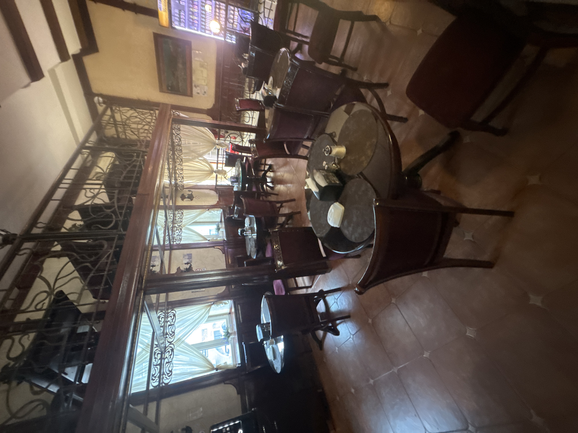 The image depicts the interior of a restaurant in Rostov-on-Don, Russia. The scene features multiple dining tables and chairs arranged throughout the space. The decor suggests a traditional or rustic ambiance, with dark wood furnishings and ornate metal railings on a mezzanine level. Large windows with sheer curtains are visible, allowing natural light to enter the room. On some tables, there are place settings with napkins and condiments. The floor is tiled. A display shelf with bottles and a glowing light fixture is visible in the background. The lighting in the restaurant is warm and inviting. There are no people present in the image.