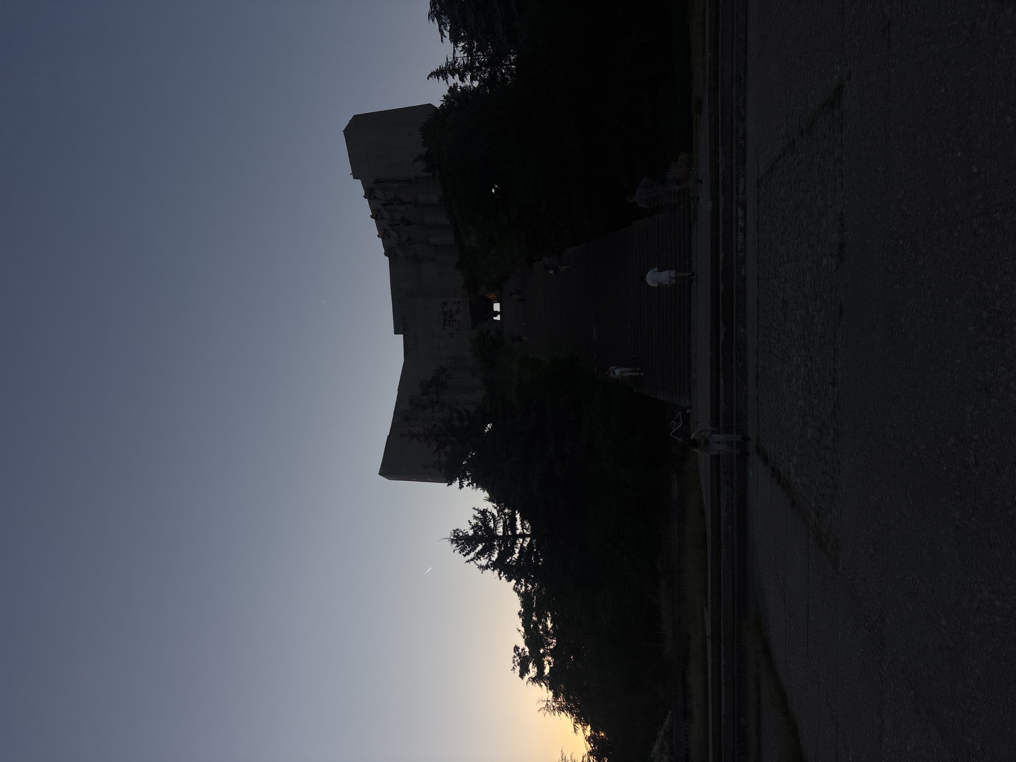 The image shows the Monument "1300 Years of Bulgaria" in Varna, Bulgaria, at dusk. A large, imposing concrete structure, resembling an open book or wings, is perched atop a hill, silhouetted against a twilight sky. A long, wide set of stairs ascends the hill towards the monument, with a few people visible on them, some walking up, others standing. The surrounding area is densely wooded with dark trees, and the foreground shows a paved area and more stairs. The sky is a gradient of deep blue at the top, fading to a pale orange and yellow on the horizon where the sun has recently set. A faint contrail from an aircraft is visible in the upper left portion of the sky. The overall scene evokes a sense of quiet reverence and the fading light of day.