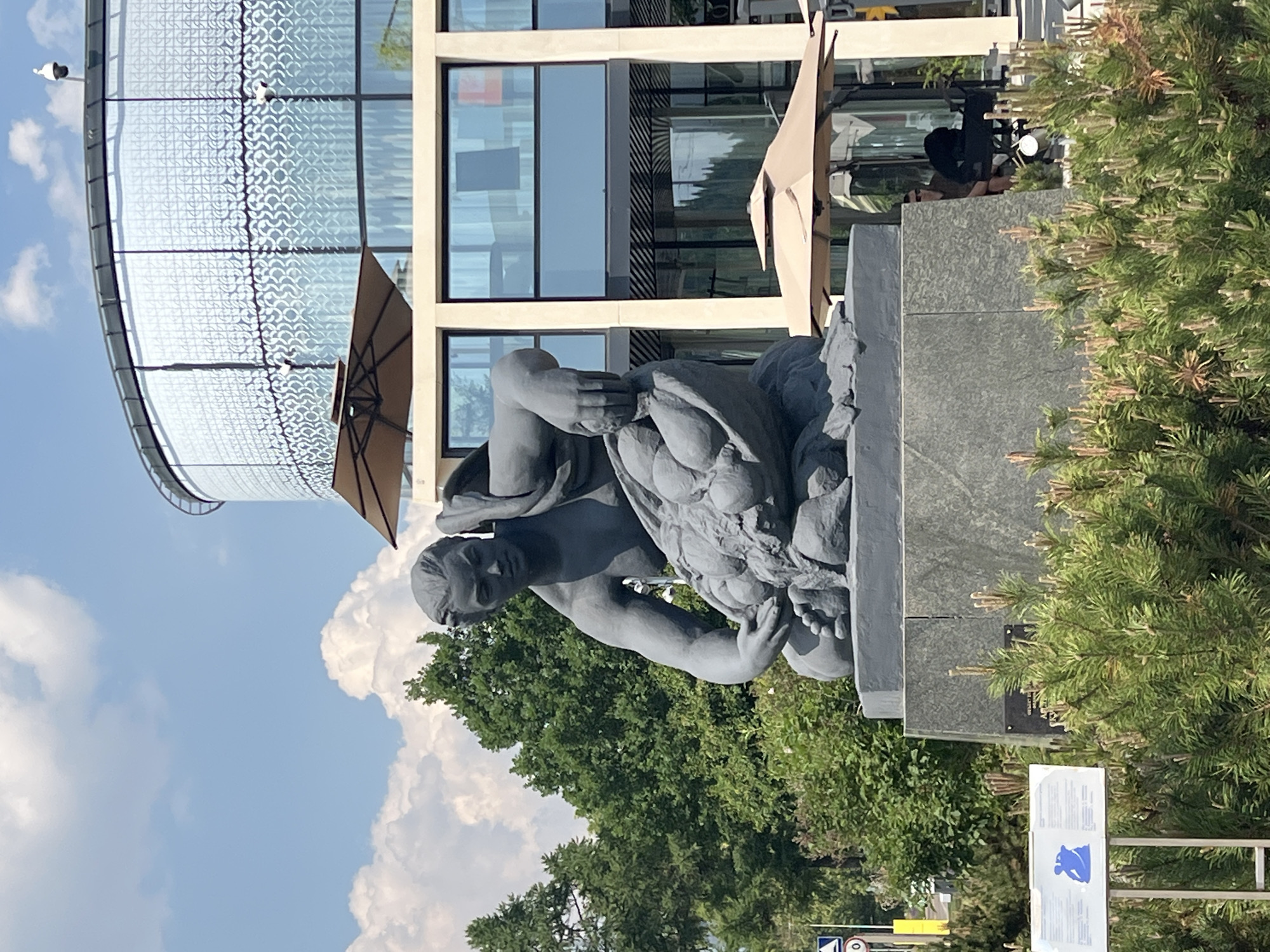 A gray stone sculpture of a seated, muscular figure is positioned in front of a modern building. The sculpture depicts a person with their legs bent and one arm reaching down to a large bag filled with round objects, possibly fruits or vegetables. The figure is draped with a piece of cloth over their shoulder and chest. The sculpture rests on a dark gray stone pedestal, which is itself placed on a larger, multi-leveled dark gray base.
Behind the sculpture, a contemporary building with large glass windows and a curved facade is visible. The windows have a decorative pattern, and several outdoor umbrellas are set up on a patio area in front of the building. Green trees and lush bushes surround the sculpture and the building, suggesting an outdoor public space. The sky is blue with scattered white clouds. In the foreground, a small informational sign with blue graphics and text is placed among the green foliage. In the background, a traffic sign with a circle and number 20 is visible. A person is seated on a chair behind some bushes near the building. The scene appears to be in an urban setting on a sunny day. This sculpture is located in Moscow, Russia, specifically near the National Research University Higher School of Economics, at the intersection of Myasnitskaya Street and Balyashniy Lane.