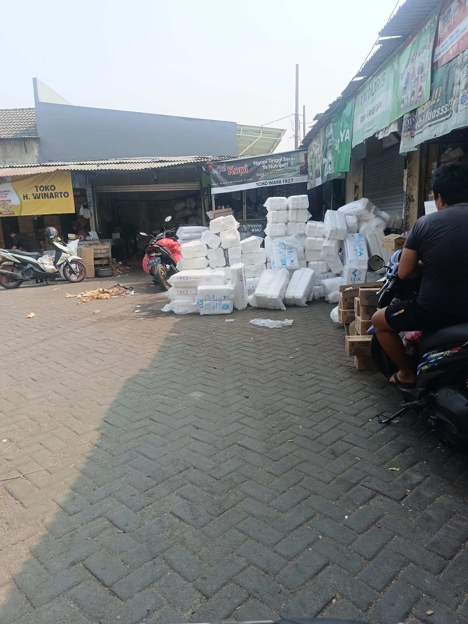 In Ketajen, Indonesia, a street market scene unfolds under a hazy sky. The foreground features a brick-paved walkway, with a motorcycle parked on the right, its rider facing away from the camera. Piles of white, boxed goods, possibly disposable containers or hygiene products, are stacked on either side of the walkway, some bearing labels such as "KCS" and "LUX."
In the mid-ground, a white and a red motorcycle are parked. Behind them, a row of market stalls with corrugated metal roofs is visible. One stall displays a yellow banner with "TOKO H. WINARTO" written in black text. Another stall features green banners with Indonesian text, including "NGKOPI TINGGI SERAT, YA NUTRIJELL!" and "TOKO WANI 1927." Inside the stalls, shadowy figures and more stacked goods are discernible. The overall impression is a bustling commercial area with goods being sold and displayed.