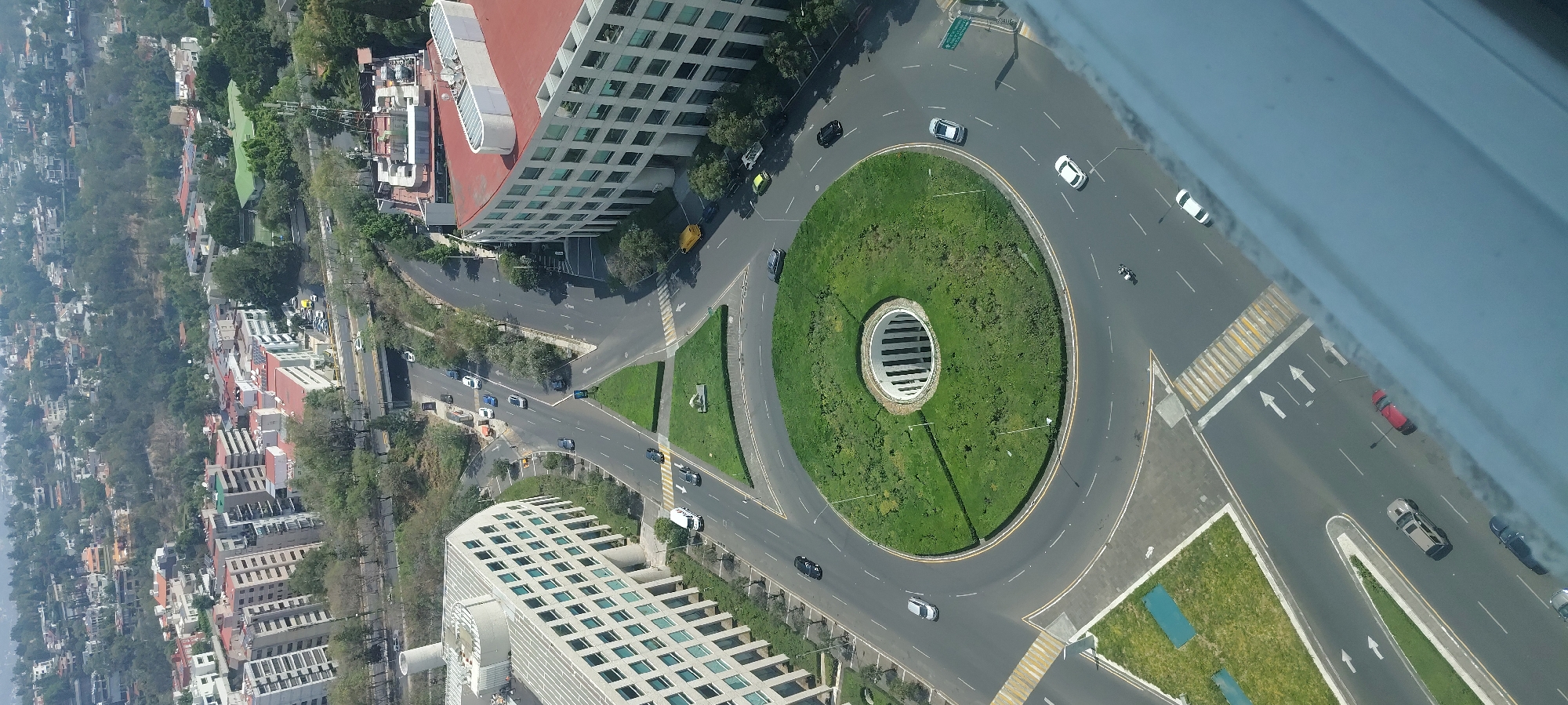 This is an aerial view of a roundabout in Mexico City, Mexico. The roundabout is circular with a green space in the center, including a large cylindrical structure with horizontal slats. There are several roads leading into the roundabout, with cars and at least one motorcycle visible on the roads. Buildings with multiple windows are on opposite sides of the roundabout. There are also trees and other greenery around the buildings and along the roads.