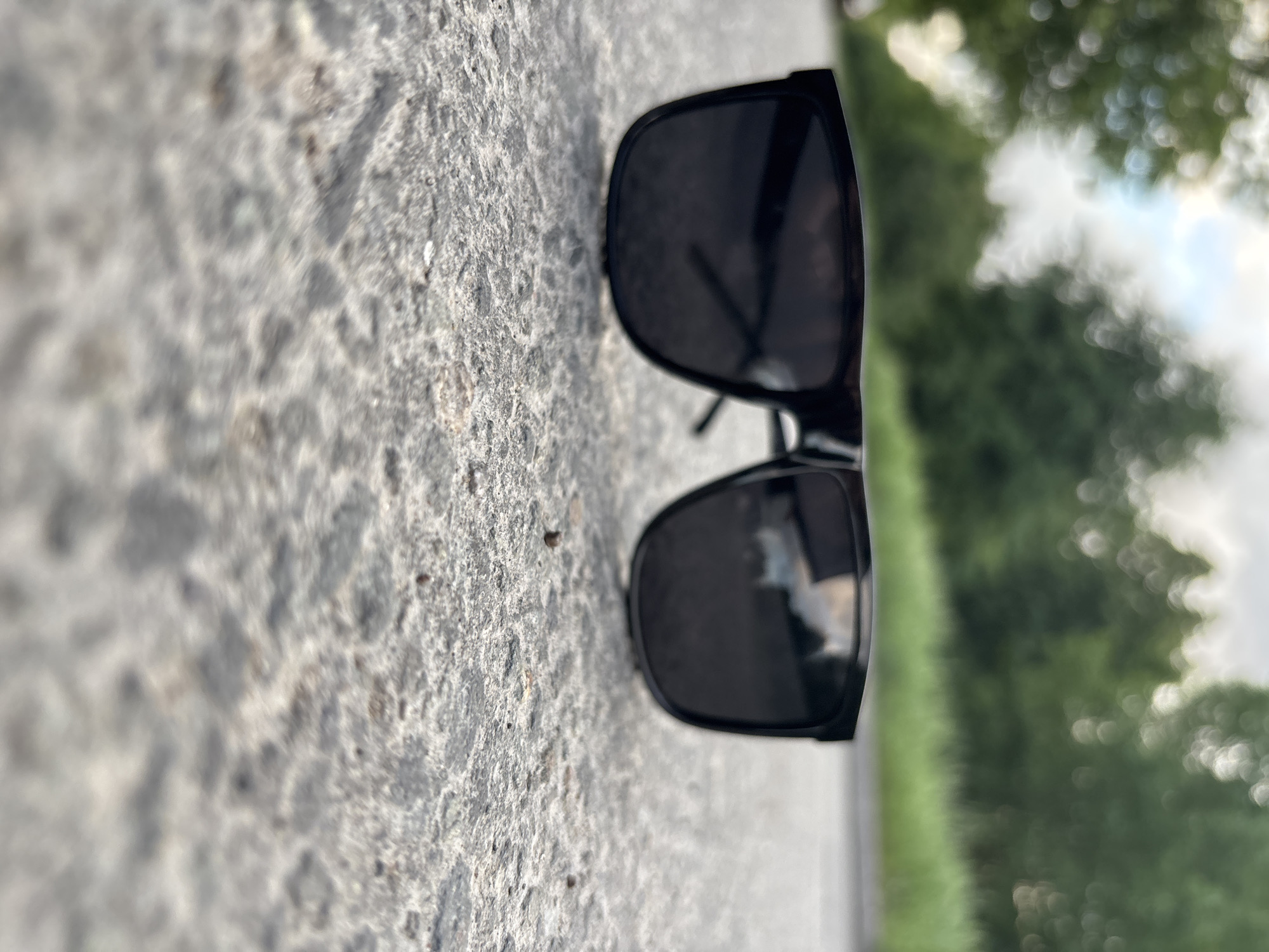 A pair of black sunglasses lie on a concrete surface, seemingly a sidewalk or road, with a blurred background of green foliage, possibly trees, suggesting an outdoor setting within the city of Unknown, Unknown. The glasses are oriented lengthwise across the image, casting a slight shadow, and reflecting the surrounding environment. The concrete surface exhibits a rough texture.