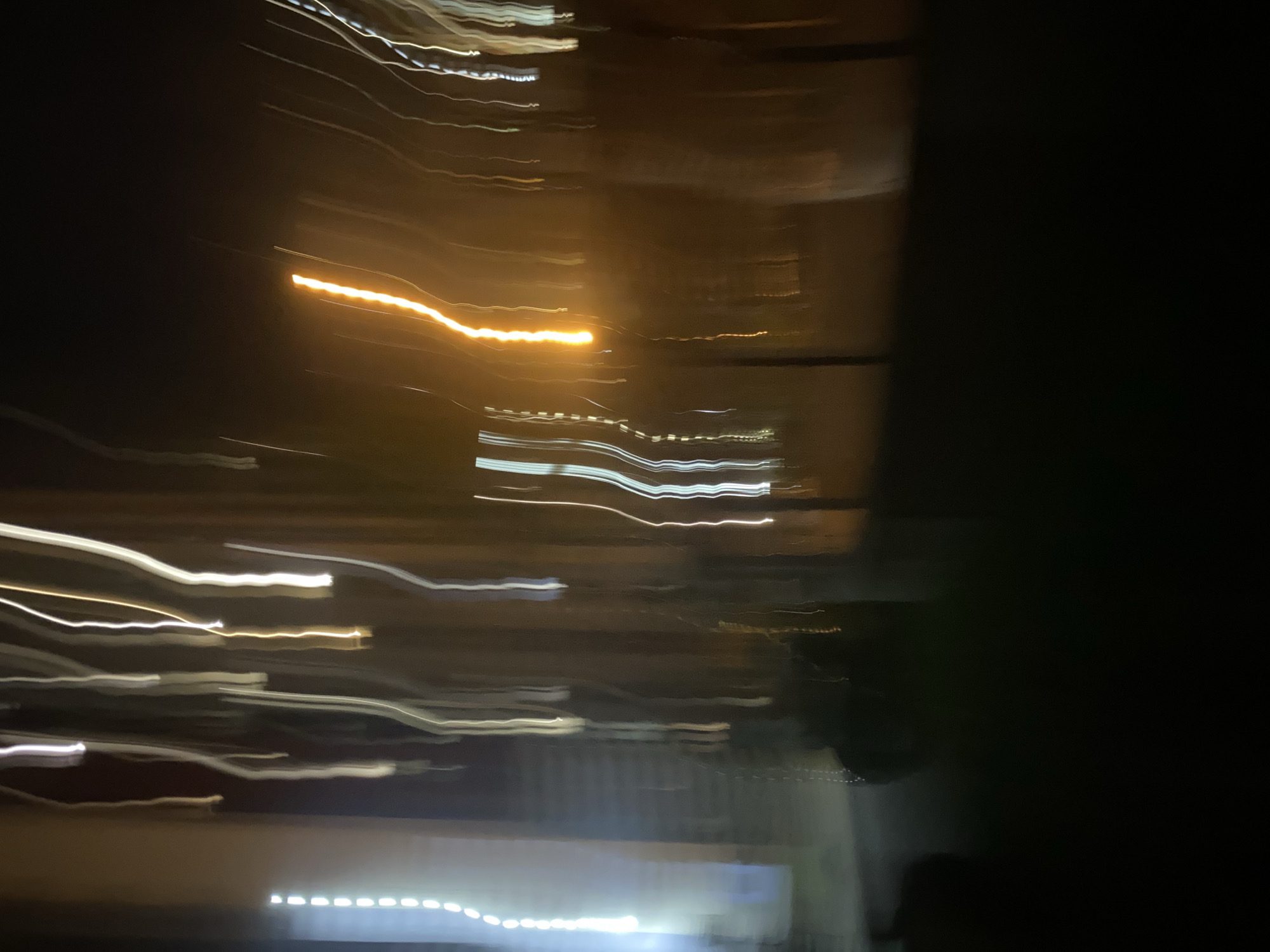 The image is a long exposure photograph taken at night, capturing light trails. The scene appears to be an outdoor urban environment, possibly a street or walkway, with buildings visible in the background. There are distinct vertical streaks of light, predominantly white and yellow, indicating artificial light sources such as streetlights or illuminated windows. Some of these light trails are sharp and defined, while others are blurred, suggesting movement during the exposure. The foreground is dark, with a shadow-like shape in the lower left quadrant, possibly a person or an object. The overall impression is one of motion and the abstract beauty of light in darkness. The location is specified as Kyiv, Ukraine, which suggests the context of the cityscape during the nighttime. There are no specific landmarks or identifiable structures that can pinpoint an exact location within Kyiv. The photograph likely captures a moment of nocturnal urban activity, with the motion blur creating a dynamic and artistic effect.