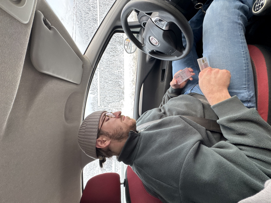 An adult male is seated in the driver's seat of a car, identified as a Fiat by the steering wheel emblem. He wears a grey ribbed beanie, round-rimmed glasses, a dark grey fleece quarter-zip pullover, and blue denim jeans. His dark hair is tied back in a small bun, and he has a short beard. He is secured by a seatbelt across his chest and lap. His gaze is directed downwards towards his hands and lap. In his left hand, he holds a smartphone enclosed in a transparent case. His right hand holds a small, rectangular card, resembling an identification or driver's license, with a second identical card resting on his right thigh. The car's interior features a light grey headliner, a black dashboard, and red-accented seats. Visible through the car's windows is a snowy, tree-lined landscape, indicative of a winter environment. The precise location within Tutin Municipality, Serbia, cannot be determined from the visual information provided, but the outdoor setting suggests a rural or mountainous area.