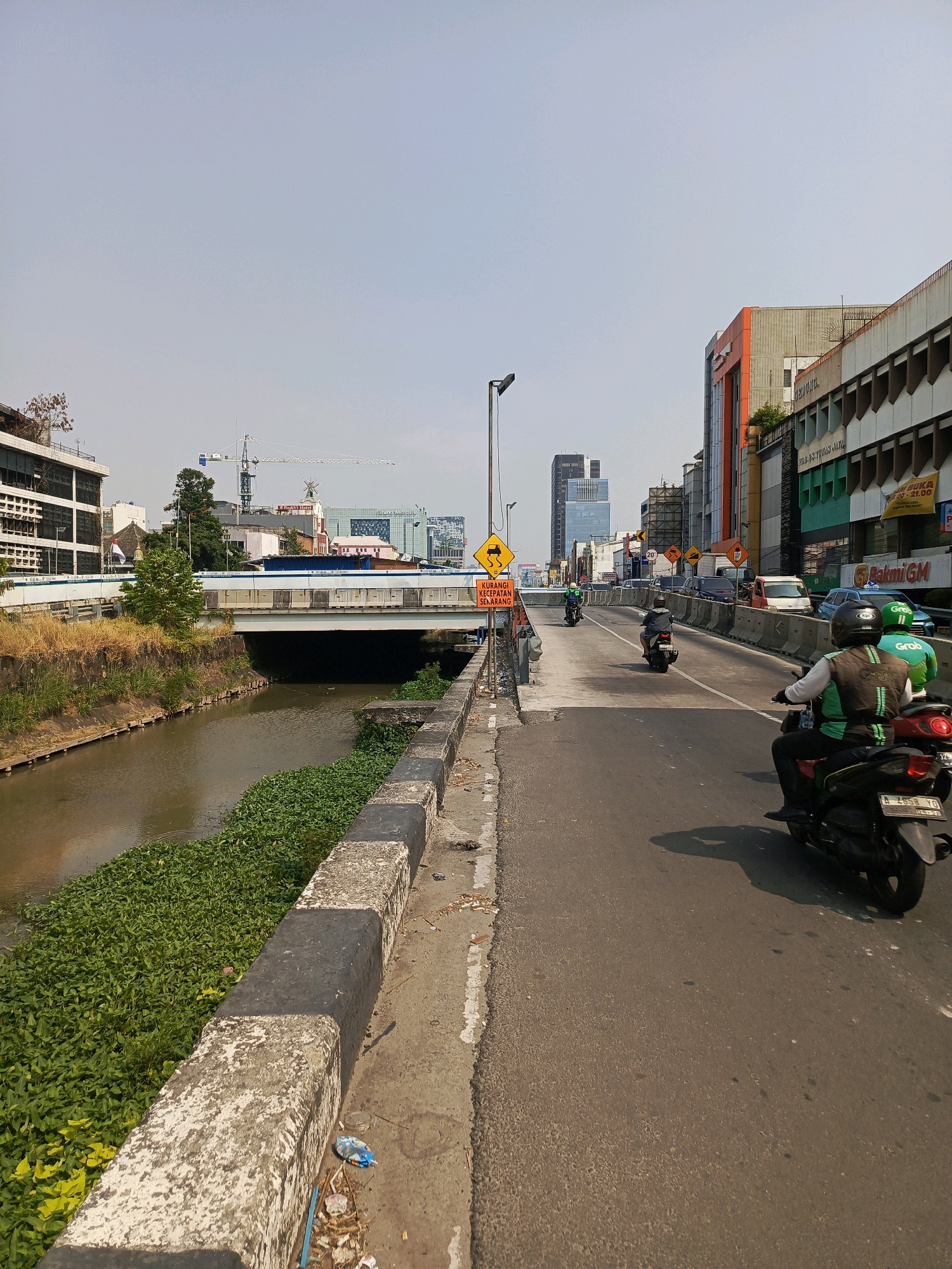The image depicts a street scene in Central Jakarta, Indonesia, with a canal running alongside the road. The canal is filled with murky water and overgrown green vegetation. The road is asphalt and appears to be a busy thoroughfare, with several motorcycles and cars visible.
In the foreground, a person on a black motorcycle wearing a green and brown vest, likely a ride-sharing service driver, is heading away from the camera. Behind them, another motorcycle with a driver in a similar uniform is also traveling in the same direction. A third motorcycle is further up the road, and several cars are on the opposite side of the road, moving in the same direction.
The roadside features a concrete barrier separating the road from the canal. Beyond the canal, there are buildings of varying heights and architectural styles, including some modern high-rise structures and older, lower buildings. A bridge spans the canal in the mid-ground. Traffic signs are present along the road, including a yellow warning sign for slippery conditions. Construction cranes are visible in the background, suggesting ongoing development in the area. The sky is clear and bright, indicating a sunny day. The overall impression is a bustling urban environment with a mix of residential, commercial, and infrastructure elements.