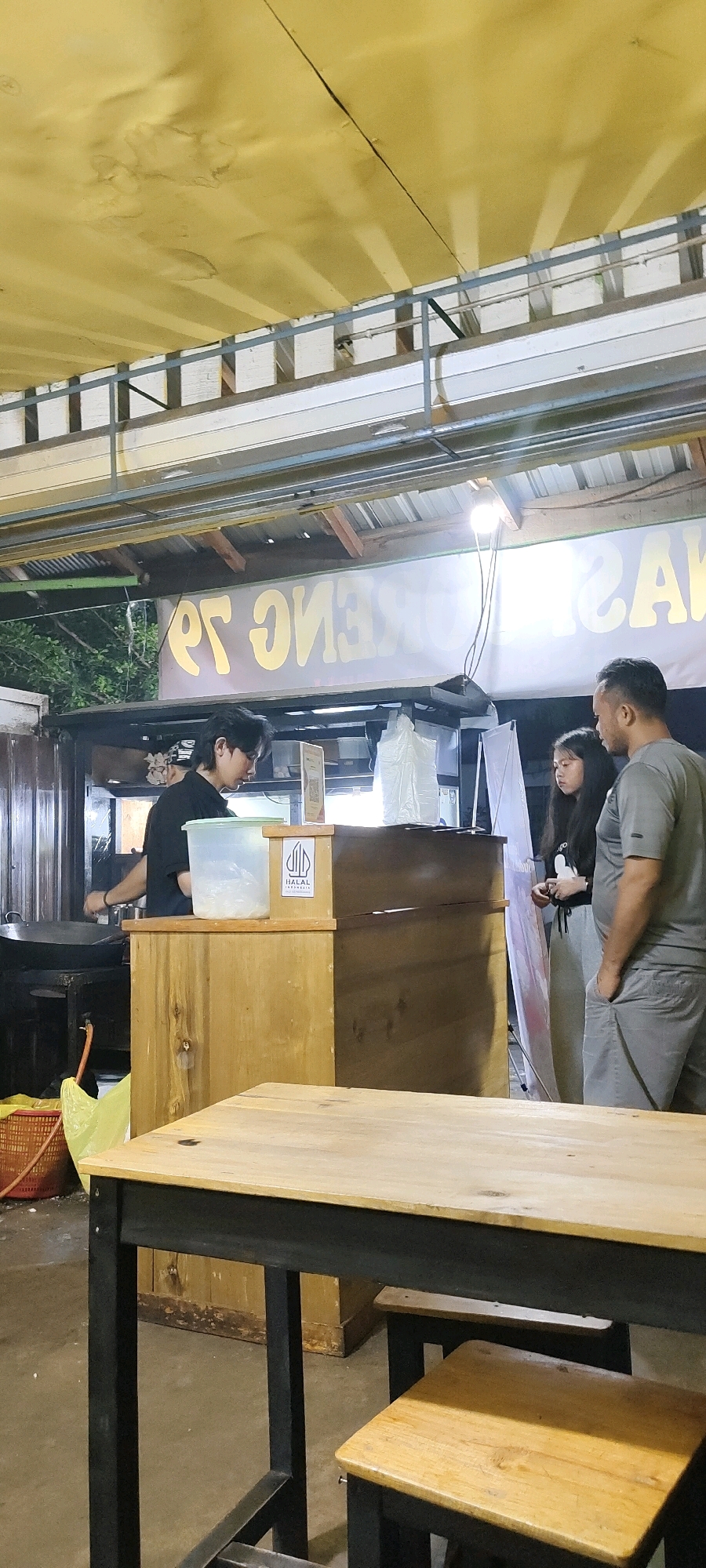 The image captures a night scene at a street food stall in Palangka Raya, Indonesia. Three individuals are present: a young man with dark hair, wearing a black shirt, is engaged in food preparation behind a wooden counter. To his right, a young woman with long dark hair, wearing grey sweatpants and a t-shirt with a graphic print, stands next to a man with short dark hair, wearing a grey t-shirt and shorts. They appear to be waiting for or interacting with the food stall worker.
The stall itself features a wooden counter with a "HALAL INDONESIA" logo displayed prominently. Behind the counter, there are various food preparation items, including a large pot, plastic containers, and bags. A sign with large yellow lettering, which appears to be in Indonesian, is visible in the background, though the text is partially obscured and reversed. The environment is covered by a yellow awning supported by metal structures, and lighting fixtures are visible overhead, indicating it's an outdoor setting. In the foreground, a wooden table with black metal legs and a wooden stool are set up, suggesting a seating area for customers. The ground is paved and appears slightly wet. The overall impression is of a bustling night market or roadside eatery.