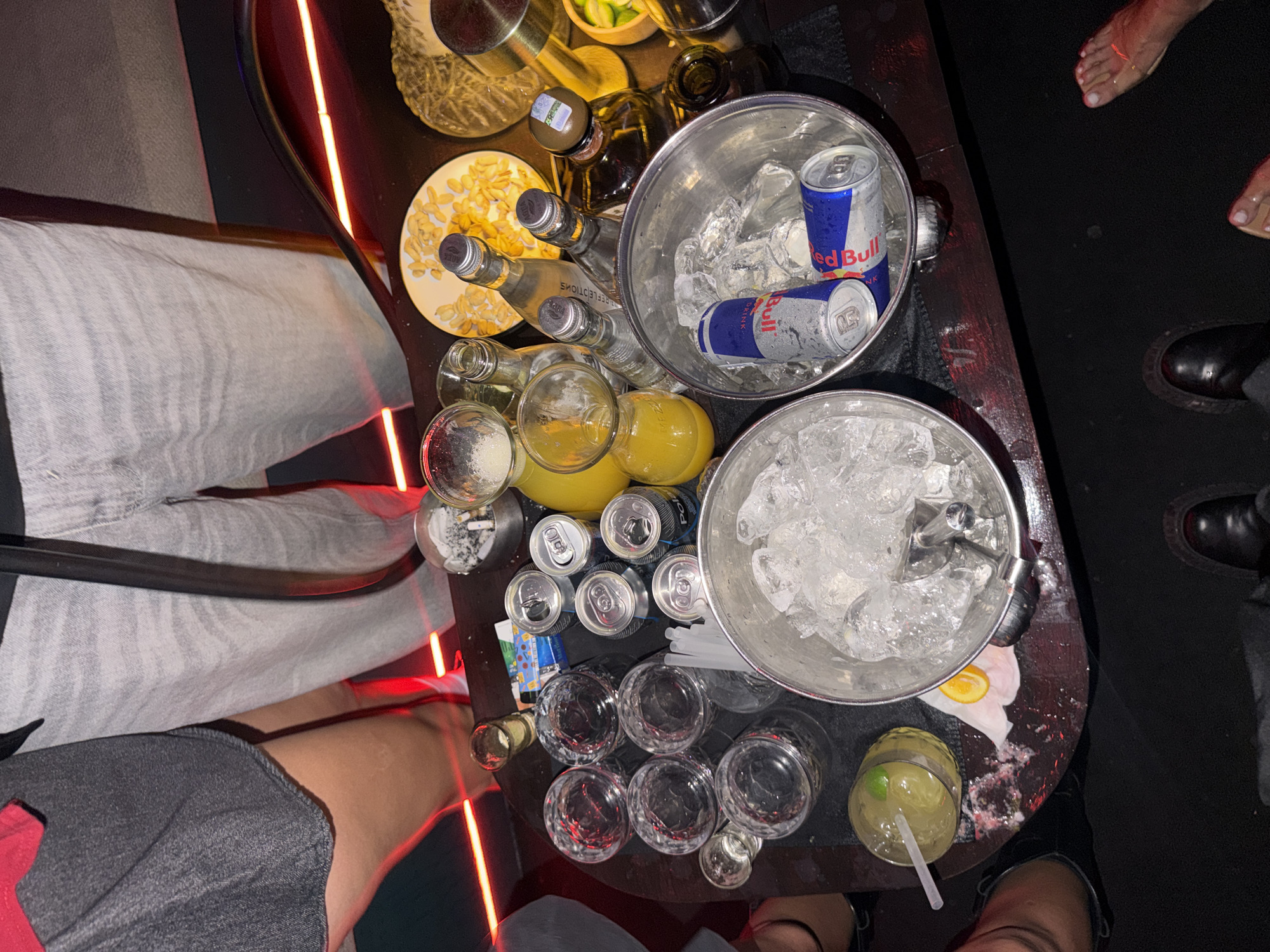 In an indoor setting with red neon lighting, a group of people are gathered around a dark table laden with drinks and snacks. In the foreground, two silver ice buckets are visible, one filled with ice and two Red Bull cans, the other containing only ice. Several glasses, some empty and some containing drinks, are arranged on the table, along with bottles of liquor and mixers. A plate of what appears to be crackers or chips sits near a bottle of clear liquid. To the left, a person's legs are visible, wearing a grey skirt and a red top. To the right, another person's feet are shown, bare and adorned with what looks like a gold anklet. The overall impression is that of a lively gathering, possibly a party or a night out.