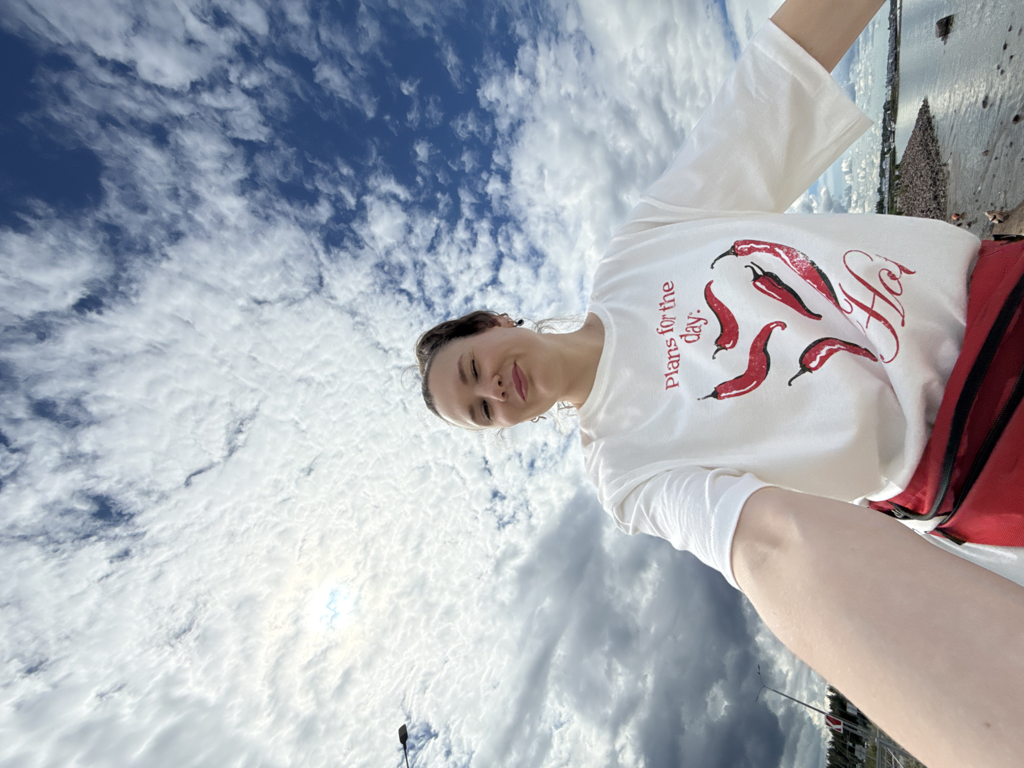 This is a low-angle, self-taken photograph of a young woman outdoors. She is wearing a white t-shirt with a graphic design of red chili peppers and the text "Plans for the day: Ho". She has her hair pulled back and is smiling. A red bag with black straps is visible at her side. The background is dominated by a dramatic sky filled with white and grey clouds against a deep blue sky. The sun is partially visible, suggesting it might be daytime, possibly late afternoon or early evening given the dramatic cloud formations. The woman is positioned in the foreground, with the sky filling the majority of the frame. In the distant background, there appears to be a body of water, a shoreline with some structures, and a large, cone-shaped object which might be a landmark or artwork. There are also hints of infrastructure like streetlights and traffic signs, suggesting an urban or semi-urban environment. The overall mood of the photo is casual and cheerful, captured against a striking natural backdrop. Based on the location context provided, this scene is likely in Saint Petersburg, Russia.