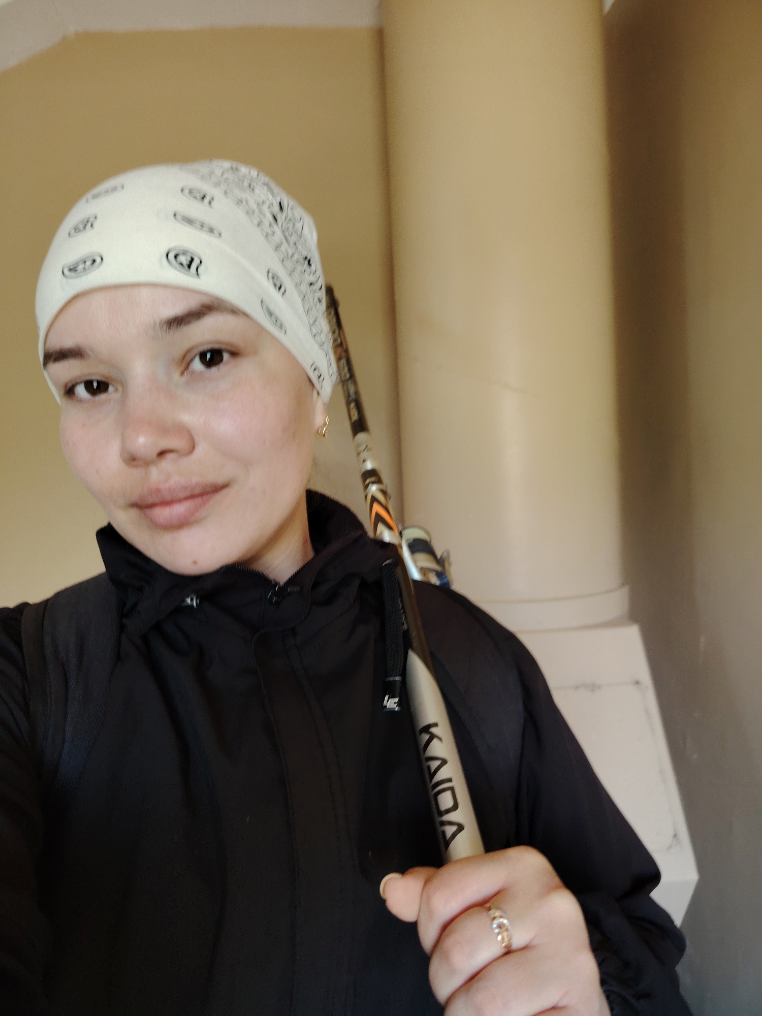 A waist-up shot shows a woman with brown eyes and fair skin wearing a patterned white bandana, a black zip-up jacket, and carrying a hockey stick. The stick, partially visible, bears the inscription "KAIDA." She is wearing a ring on her finger. The background consists of a beige-toned wall with a visible cylindrical column and a white, step-like structure at its base. The location is Марковский, Russia.