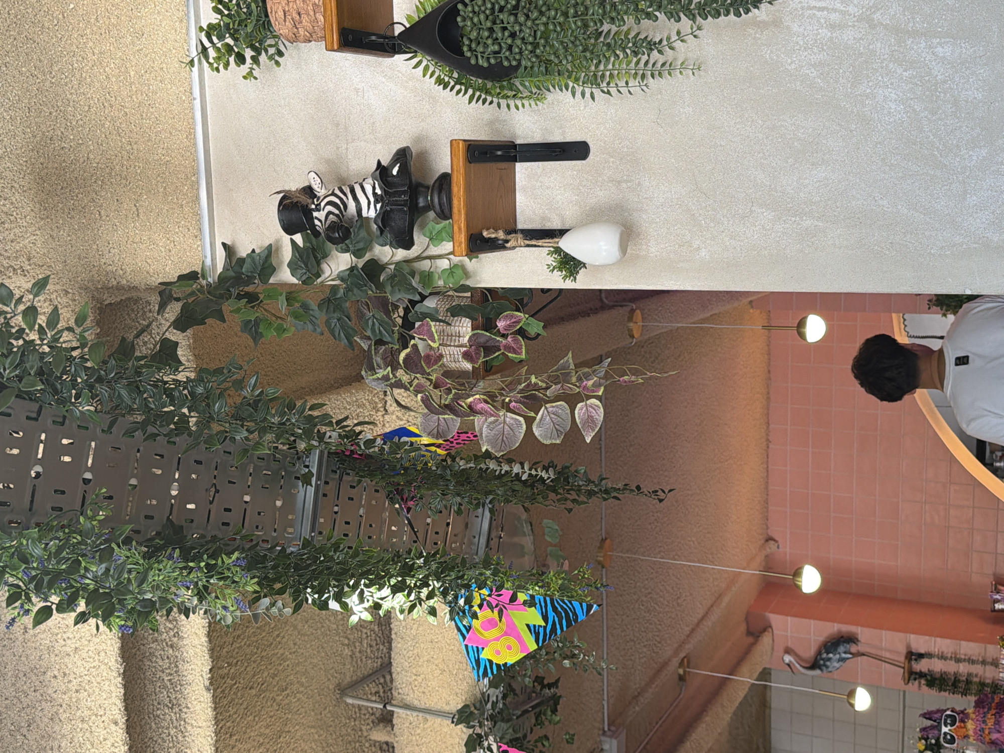 This image shows an indoor setting, possibly a cafe or shop, decorated with lush greenery and whimsical accents. The scene is brightly lit, suggesting daytime. In the foreground, a person wearing a white t-shirt is partially visible, looking away from the camera.
The decor features several interesting objects. A decorative zebra head is mounted on a wooden plank, beside a white, egg-shaped object hanging from a rope. Further down, a decorative flamingo statue stands near a display of colorful flags or banners with geometric patterns. The walls are textured, with one section appearing to be tiled in a soft pink color. The overall ambiance is playful and eclectic, with a strong emphasis on plants and unique decorative items. The presence of cascading plants and artificial greenery adds to a vibrant and lively atmosphere.