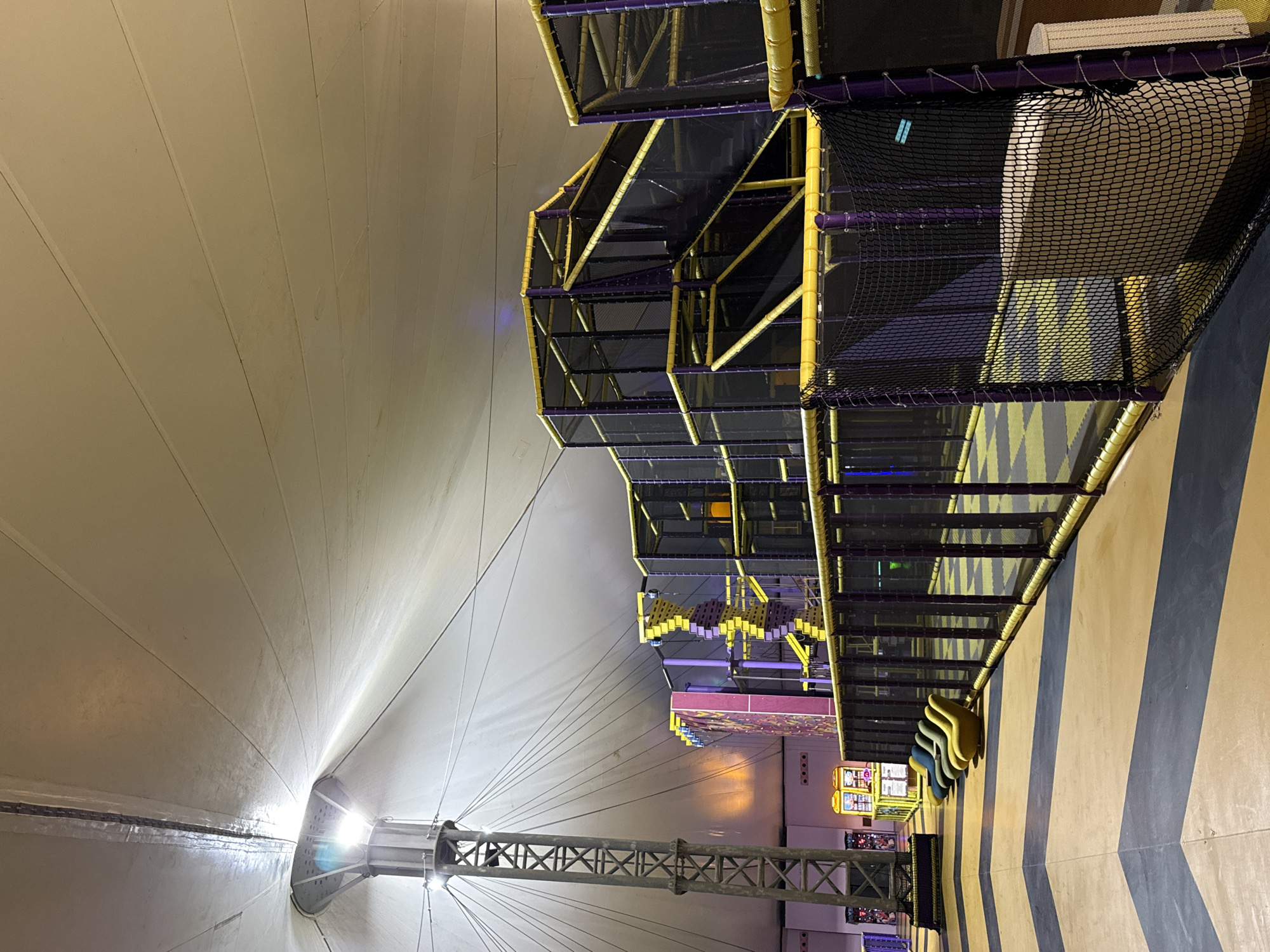 The image shows the interior of a large, tent-like structure, likely an indoor playground or activity center in Bodrum, Turkey. A tall, metal tower with lights is visible on the left, supporting the white, tensioned fabric ceiling that dominates the upper portion of the frame. Below the ceiling, a multi-level playground structure with purple posts and yellow accents, featuring climbing nets and slides, occupies the middle and right sections of the image. The playground is enclosed by black safety netting with yellow and purple trim. In the foreground on the left, several brightly colored, beanbag-like chairs are lined up on a patterned floor. The overall lighting suggests an indoor, possibly evening or dimly lit environment.