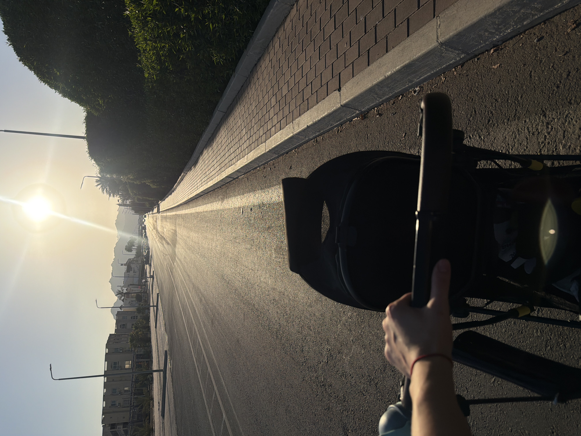 The image captures a first-person perspective, likely of someone pushing a stroller, along a paved walkway adjacent to a road. The sun is low in the sky, casting a warm, golden light and creating long shadows. The time of day is likely late afternoon or early morning, given the angle of the sun and the quality of light. The weather appears to be clear and sunny.
The scene is urban, with buildings and palm trees visible in the distance. The walkway is paved with brick-like tiles and has a concrete border. The road next to it is asphalt, with visible lane markings. A person's hand, wearing a thin red bracelet, is visible gripping the handle of what appears to be a stroller. The stroller itself is dark-colored and partially visible.
In the background, mountains are silhouetted against the bright sky, suggesting a location with a dramatic landscape. The architecture of the buildings hints at a Middle Eastern or North African setting, consistent with the provided location context of Sharm El Sheikh, Egypt. The overall mood is peaceful and serene, characterized by the gentle sunlight and the quiet scene. There is no visible text in the image.