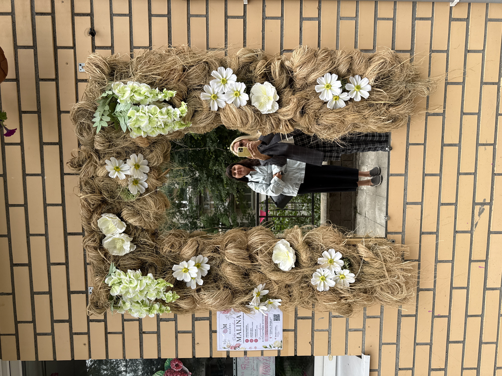 The image depicts a decorative archway made of rough twine and adorned with artificial white and light green flowers, mounted on a light brown brick wall. A rectangular mirror is integrated into the center of the archway. Two women are reflected in the mirror. One woman on the left wears a light blue denim jacket and black pants, holding her phone. The other woman on the right wears a dark blazer over a plaid skirt, also holding a phone to take a picture. Behind them, a building with windows and greenery is visible. On the left side of the brick wall, a sign with Cyrillic text and images of flowers is affixed. The sign appears to be an advertisement for "MALINA." The overall scene suggests an outdoor setting, possibly in front of a shop or establishment.