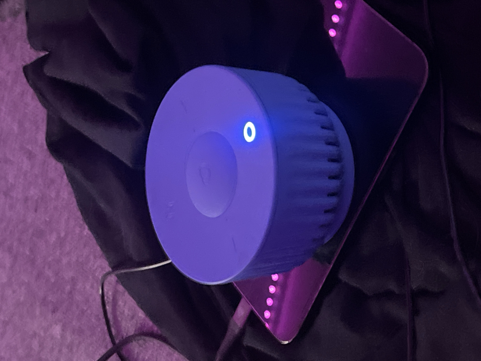 A top-down shot depicts a round, blue smart speaker with a glowing blue ring on its front face, sitting on a glossy, purplish triangular object with pink lights visible along its edges. A black cable is visible to the left of the speaker, connected to it. The background includes textured light purple carpet and a draped black fabric. There is no evidence in this image to suggest this image was taken in the city of Saint Petersburg, Russia.