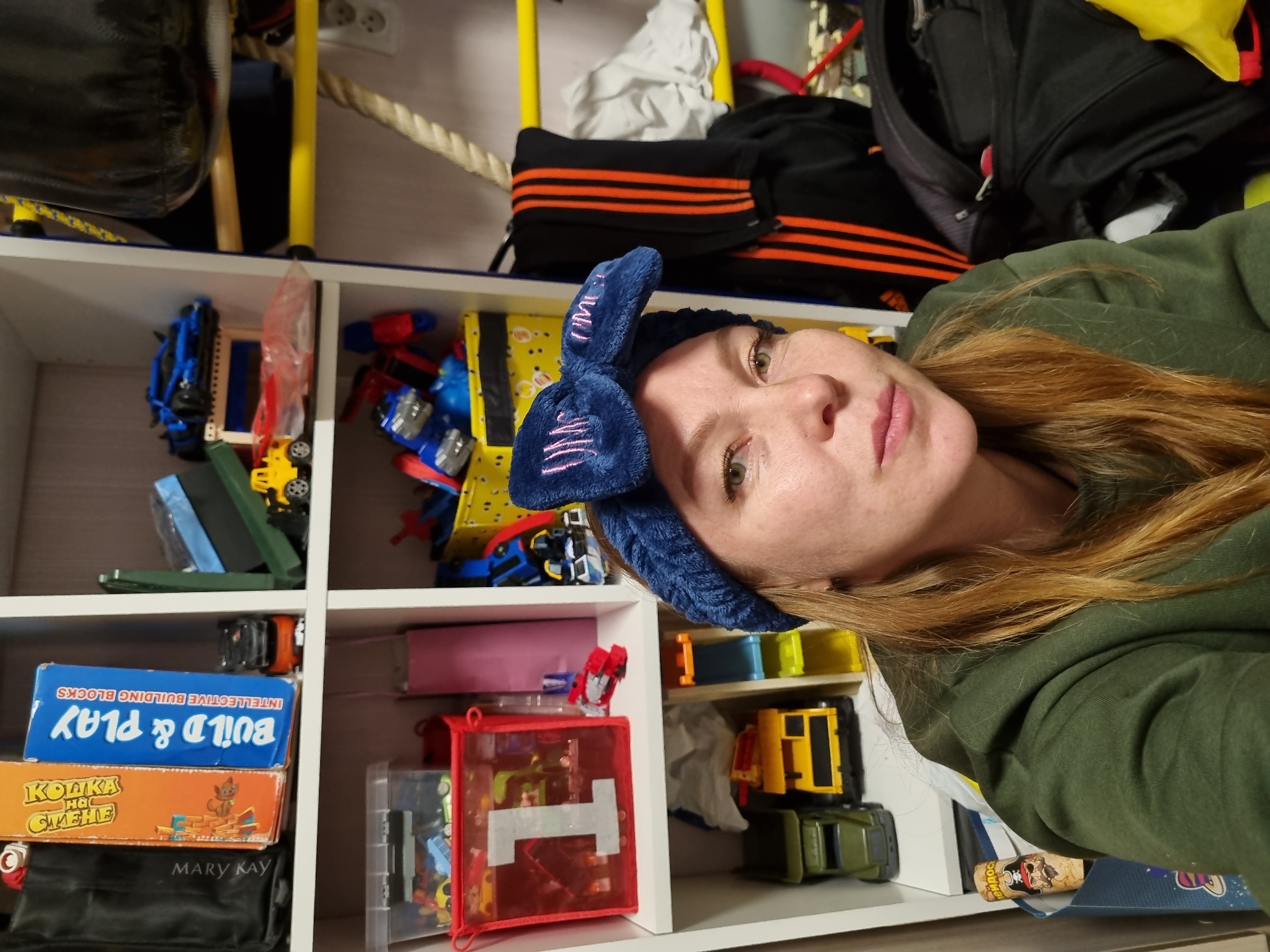 This image features a woman posing indoors, likely in a home setting in Chaikovsky, Russia. The woman's face, with fair skin, light-colored eyes, and long reddish-brown hair, is visible from her nose to her upper torso. She wears a blue fluffy headband with a bow, which has some indistinct pink stitching, and a green hooded sweatshirt. Her expression is neutral. The photograph is tilted, rotating the scene clockwise.
Behind her, a white modular shelving unit is filled with various objects, predominantly children's toys and games. On one shelf, a blue box labeled "BUILD & PLAY INTELLECTUAL BUILDING BLOCKS" is stacked on an orange box that features a fox character and the Cyrillic text "КОШКА НА СТЕНЕ" (Cat on the Wall). Below these, a black pouch with the "MARY KAY" logo is visible. Other compartments contain an assortment of toy vehicles, including cars, trucks, and possibly robots, as well as a red mesh bag with a white "T" on it.
Above the shelving unit, in the upper background, several items are stored: a black sports bag with prominent orange stripes, a black backpack, a white cloth, and parts of yellow metal bars or a frame. A wall outlet with two sockets and a thick rope are also visible on the back wall. A black, cylindrical object, possibly a punching bag, is partially seen in the upper left corner. The setting suggests a personal space, potentially a child's room or a well-used storage area within a residence.