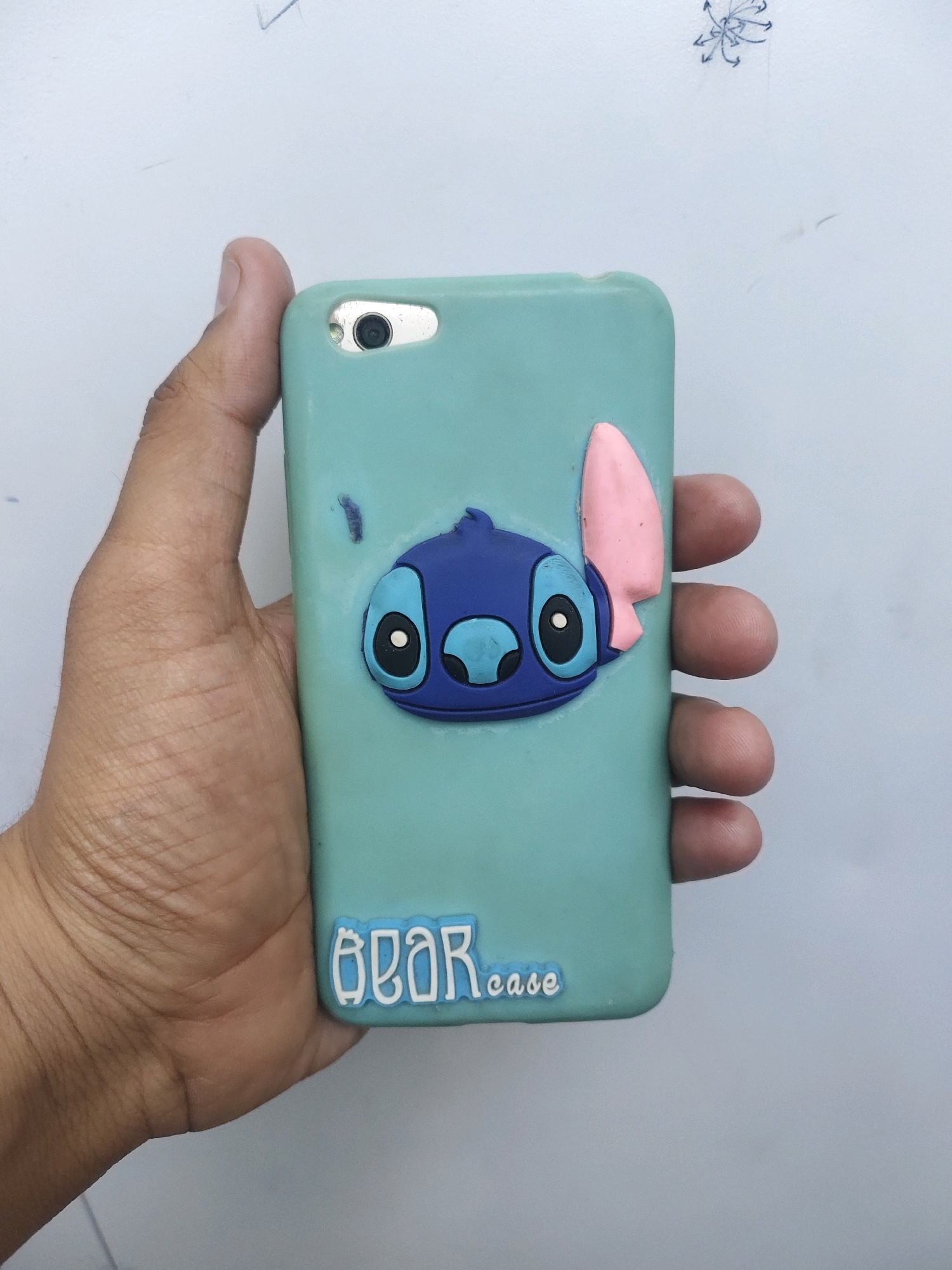 A person's hand holds a light blue phone case with a 3D Stitch character on it. The Stitch character is blue with large, round eyes and a pink ear. Below the character, the words "bear case" are written in light blue lettering. The phone case is on a white background with some faint blue markings and a small, stylized drawing at the top. The image is a close-up shot, focusing on the phone case and the hand holding it. There is no information about the location in the city of Sumerpur, India.