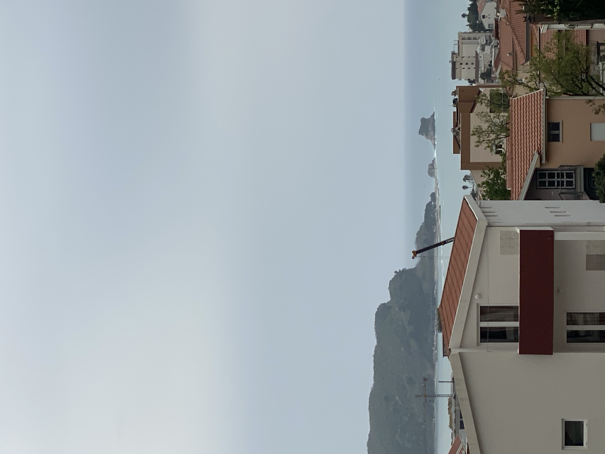 The image shows a view of the city of Budva, Montenegro, with a focus on a residential area overlooking the coastline. In the foreground, a modern white building with a red-tiled roof and a maroon vertical architectural feature is visible. The midground features a lush green mountain and a construction crane. In the distance, the Adriatic Sea stretches towards the horizon, with small islands and rock formations emerging from the water. Other buildings with red-tiled roofs are also present. The sky is overcast, creating a soft, diffused light across the scene.