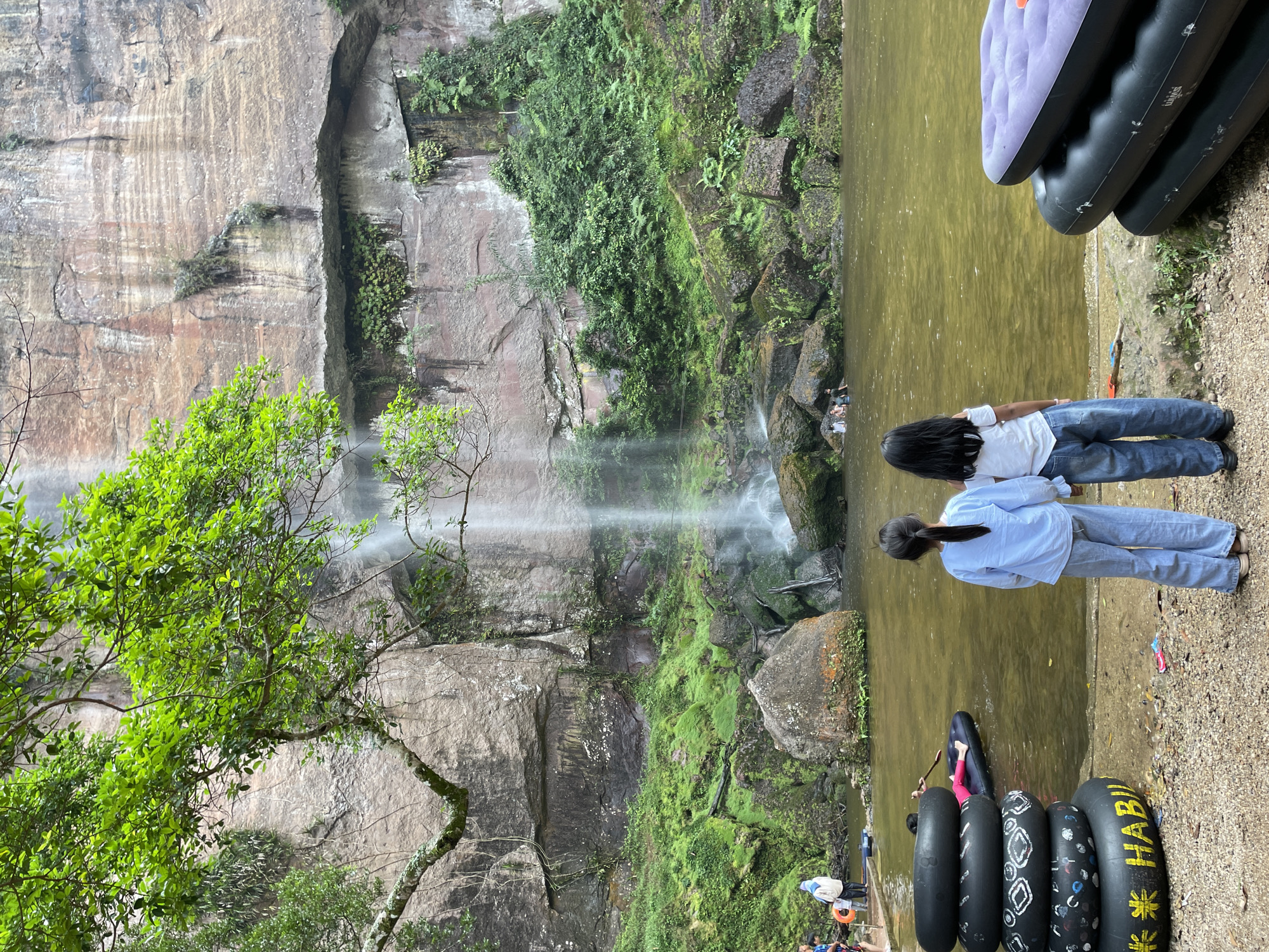 In Sarilamak, Indonesia, a waterfall cascades down a rocky cliff face, surrounded by lush green vegetation. In the foreground, two young girls stand facing the waterfall, holding hands. Several inflatable tubes are stacked on the shore to their left, and another inflatable mattress is on the right. In the water, several people are swimming and playing. The scene suggests a recreational area at the base of a waterfall.
