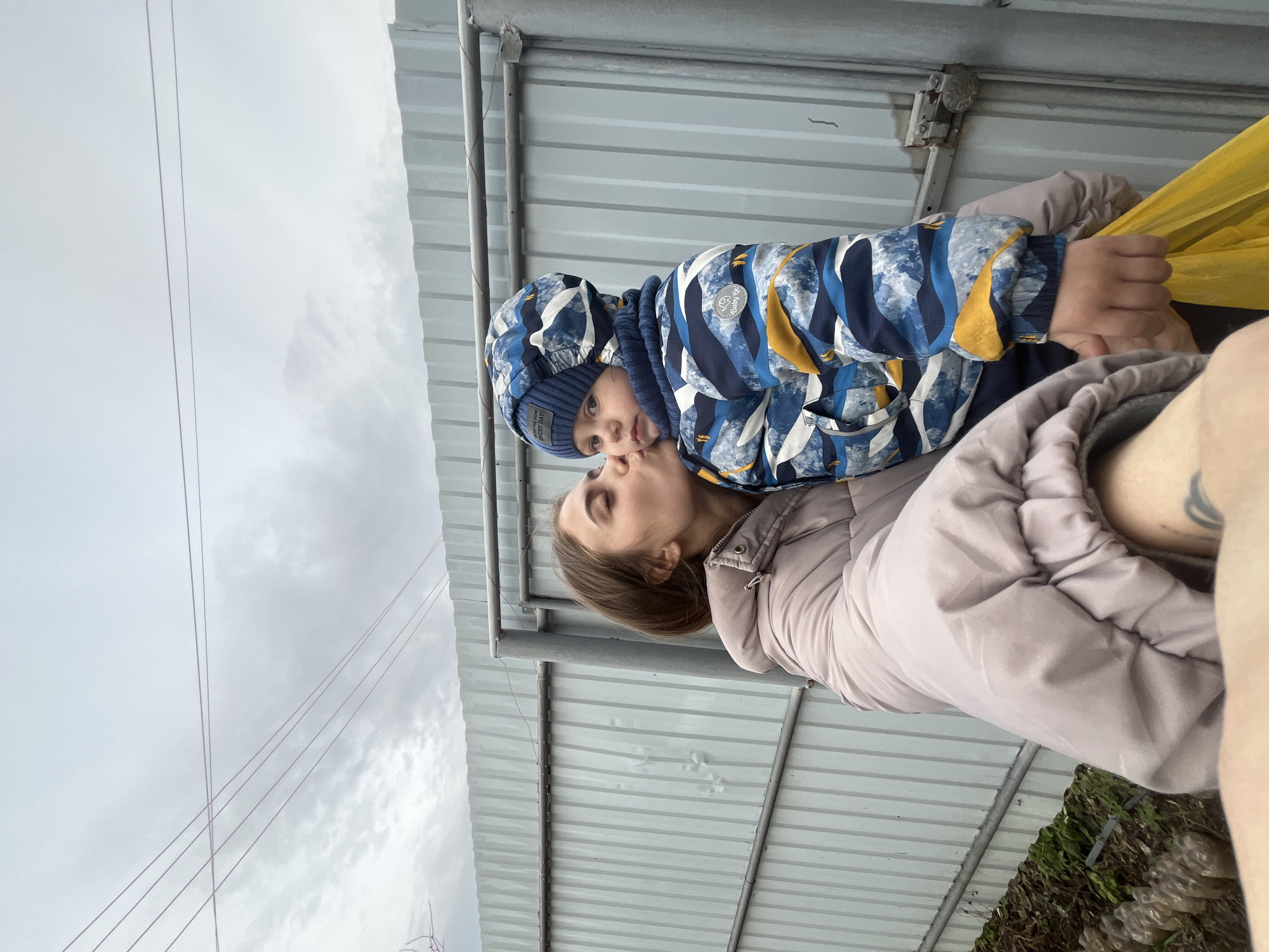The image shows a young woman holding a young child, presumably her son, in an embrace. The child is wearing a blue and yellow patterned snowsuit with a matching hat. The woman is wearing a light pink puffer jacket. They are standing underneath a corrugated metal structure, possibly a bus stop or small shelter. Outside the shelter, the sky is overcast, with power lines visible in the background. The photograph appears to be taken outdoors, possibly in the vicinity of Фоки, Russia, based on the prompt.