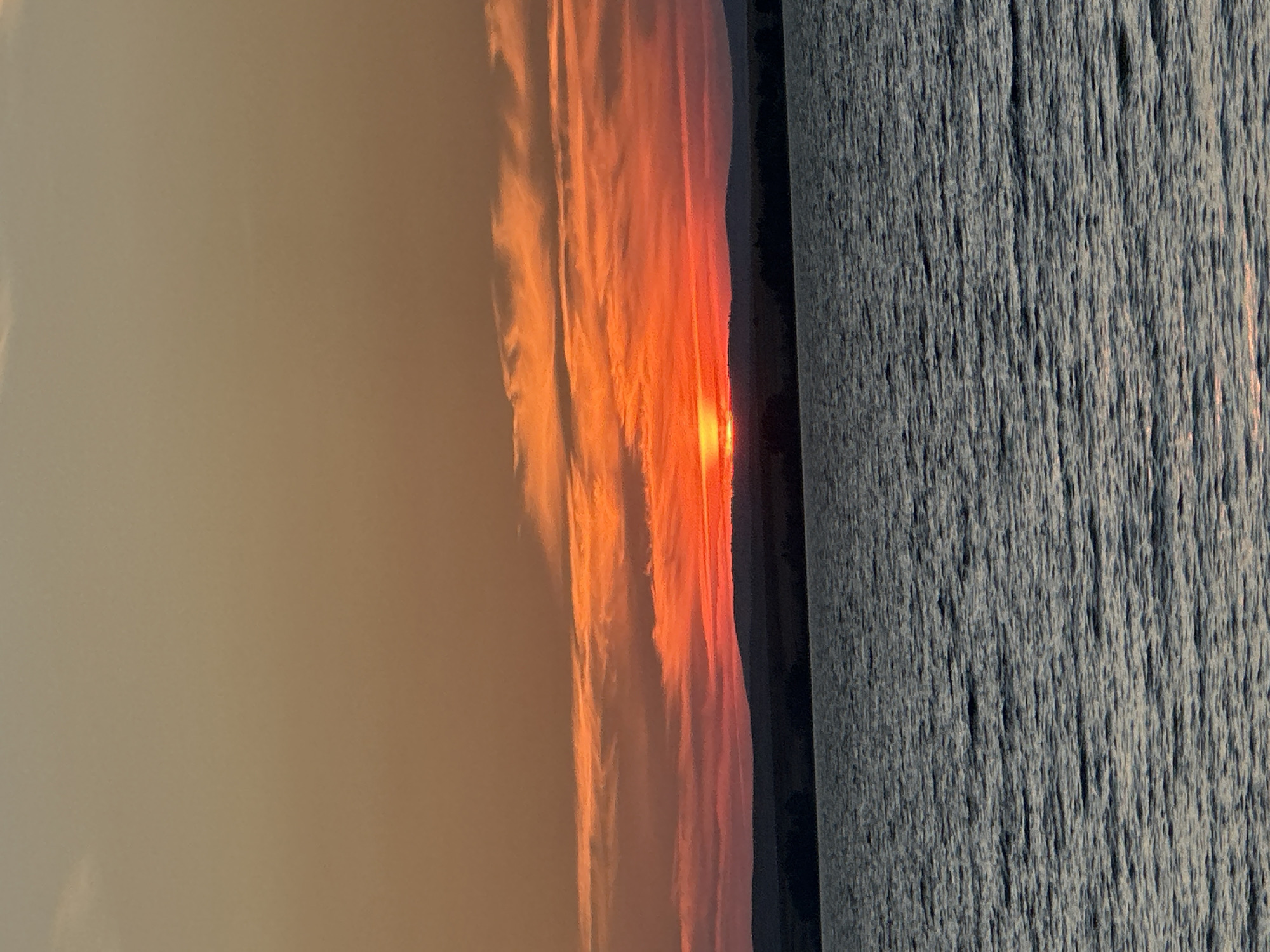 The image shows a body of water in front of a sunset. The sky is a gradient of orange and red, with streaks of clouds illuminated by the setting sun. A dark, hilly horizon line separates the sky from the water. The water's surface is rippled and reflects the colors of the sky. The scene is tranquil and appears to be an outdoor landscape, likely a lake or a large river. The location is the city of Energetik, Russia.