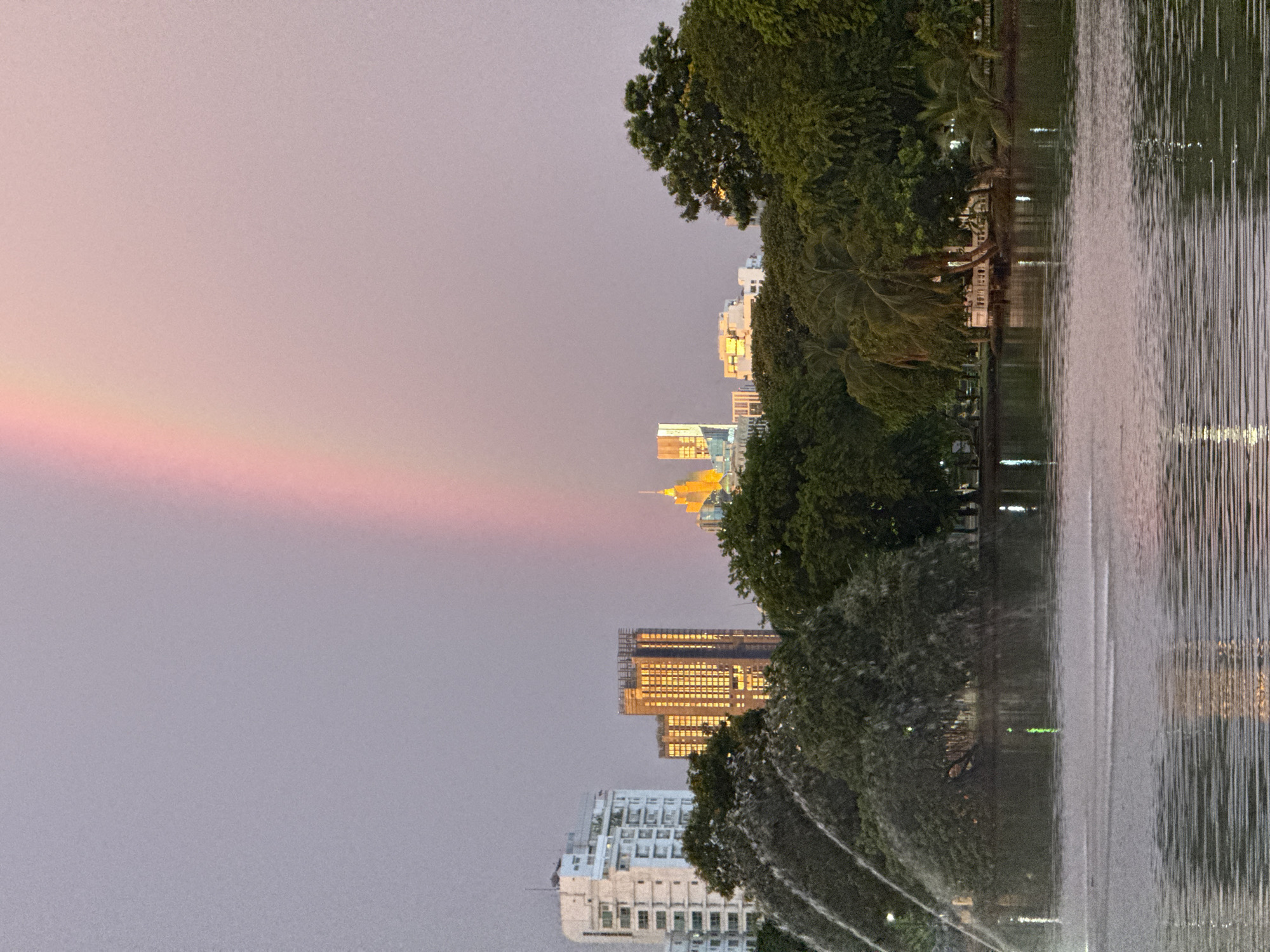 This is a photograph taken from a slightly low angle, showing a cityscape with a body of water in the foreground and a rainbow in the sky. The image is oriented vertically.
The scene depicts a body of water, likely a park lake or pond, on the right side of the frame. Water is being sprayed from fountains, creating a misty effect and a shimmering surface with reflections of lights. Lush green trees line the bank of the water on the left.
Beyond the trees, a collection of modern buildings rises, indicating an urban environment. Some buildings are brightly lit, suggesting it is either dusk or dawn. The architecture varies, with some taller skyscrapers and lower-rise structures visible. One building in particular stands out with a grid of warmly lit windows.
The sky above is a soft gradient of pink and purple, with a visible rainbow arching across the left side of the frame. The presence of the rainbow and the muted colors of the sky suggest post-rain conditions and the soft light of either sunrise or sunset.
The overall mood of the image is serene and picturesque, blending the natural beauty of the rainbow and water features with the man-made structures of the city. There is no visible text in the image. The location context provided is Bangkok, Thailand, which aligns with the presence of modern high-rise buildings and possibly a park setting.