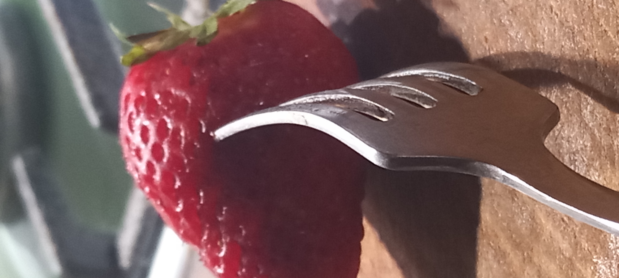 A close-up, slightly angled shot focuses on a red strawberry with its seeds visible and a green stem. A silver utensil, possibly part of a set of tongs or a decorative fork, is positioned in front of the strawberry, with its curved, flame-like tip gently touching the side of the fruit. The utensil has cut-out designs. The background is blurred, showing dark grey and lighter grey horizontal bars above a green area, possibly indicating a kitchen environment or a setting with metallic elements. The scene is lit from above, casting shadows on the wooden surface below the strawberry and utensil. There are no people or animals visible. The image does not contain any discernible elements that would place it in the city of Kryvyi Rih, Ukraine.
