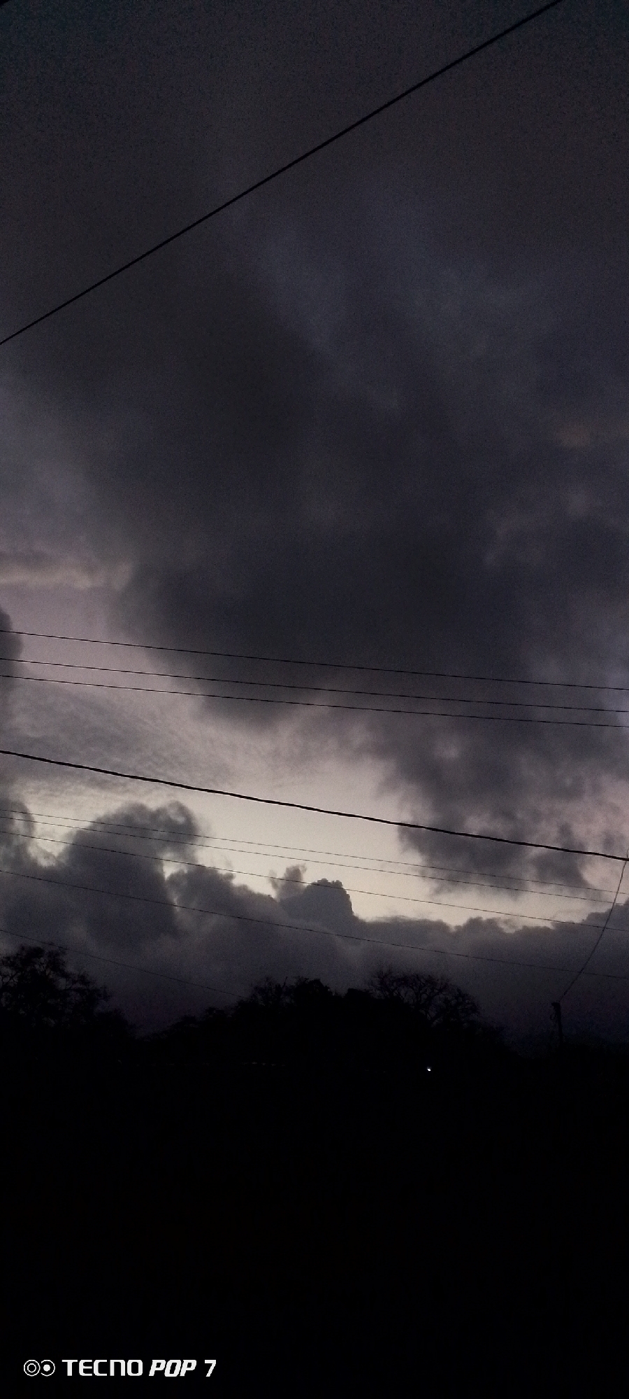 The image presents a dramatic view of a heavily overcast sky, likely at dusk or early morning, over Altagracia, Venezuela. Dark, dense grey clouds fill the majority of the frame, suggesting stormy weather or impending rain. A lighter, pale grey sliver of sky is visible near the horizon, nestled between layers of dark clouds. Several thin, dark utility or power lines stretch horizontally across the mid-ground, cutting through the sky and clouds. The bottom portion of the image is dominated by a dark silhouette of trees or dense foliage, obscuring any ground details and contributing to the somber atmosphere. No people or specific activities are visible. In the bottom left corner, the text "TECNO POP 7" is present, indicating the device used to capture the photograph.