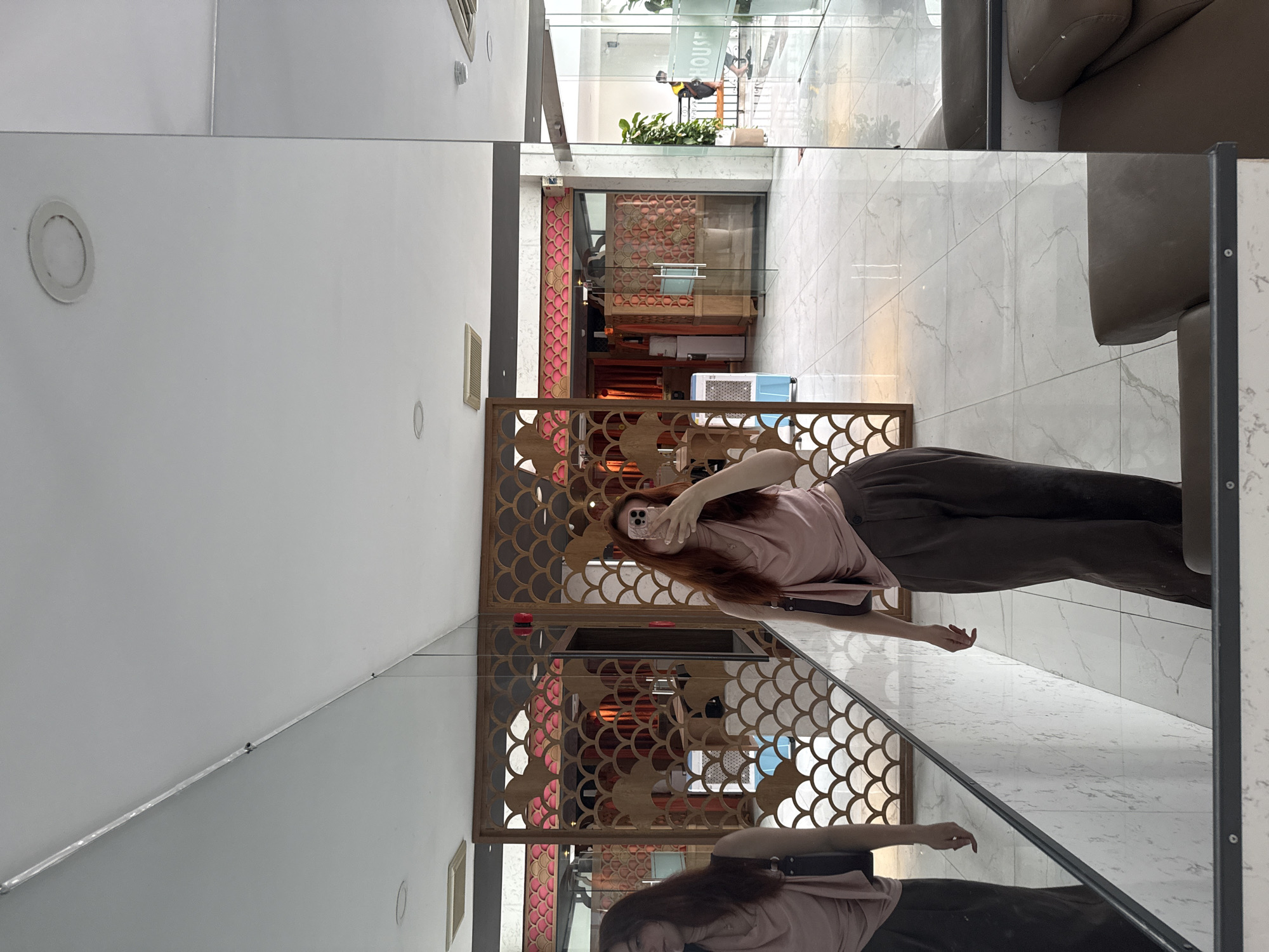 Captured in Bac Nha Trang Ward, Vietnam, the image presents a woman taking a photo of herself with a mobile phone in front of a mirror. She is wearing a light pink top and dark brown pants. The mirror is situated near a decorative lattice-style partition. The reflection also shows the interior, including tiled flooring, furniture and architectural elements.